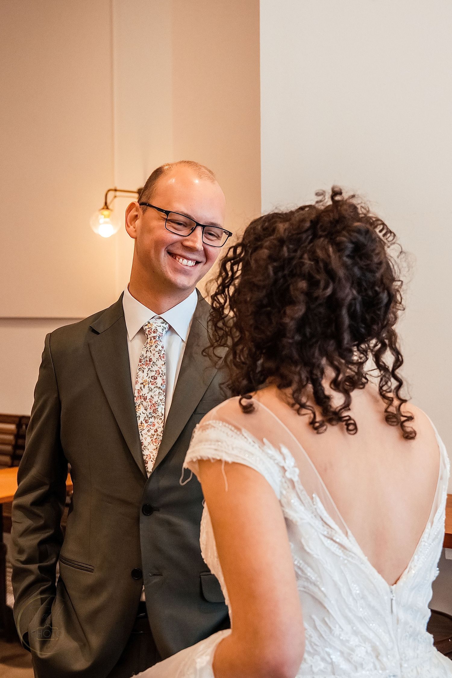 Groom in dark suit and floral tie smiling at bride in white lace dress during wedding ceremony indoors.