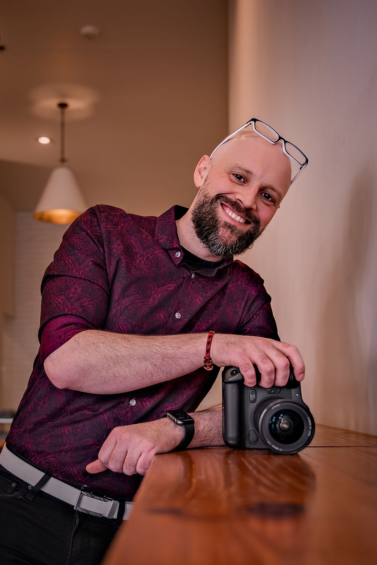 Smiling bald man with beard and glasses on head, wearing burgundy shirt, posing with camera on wooden table.
