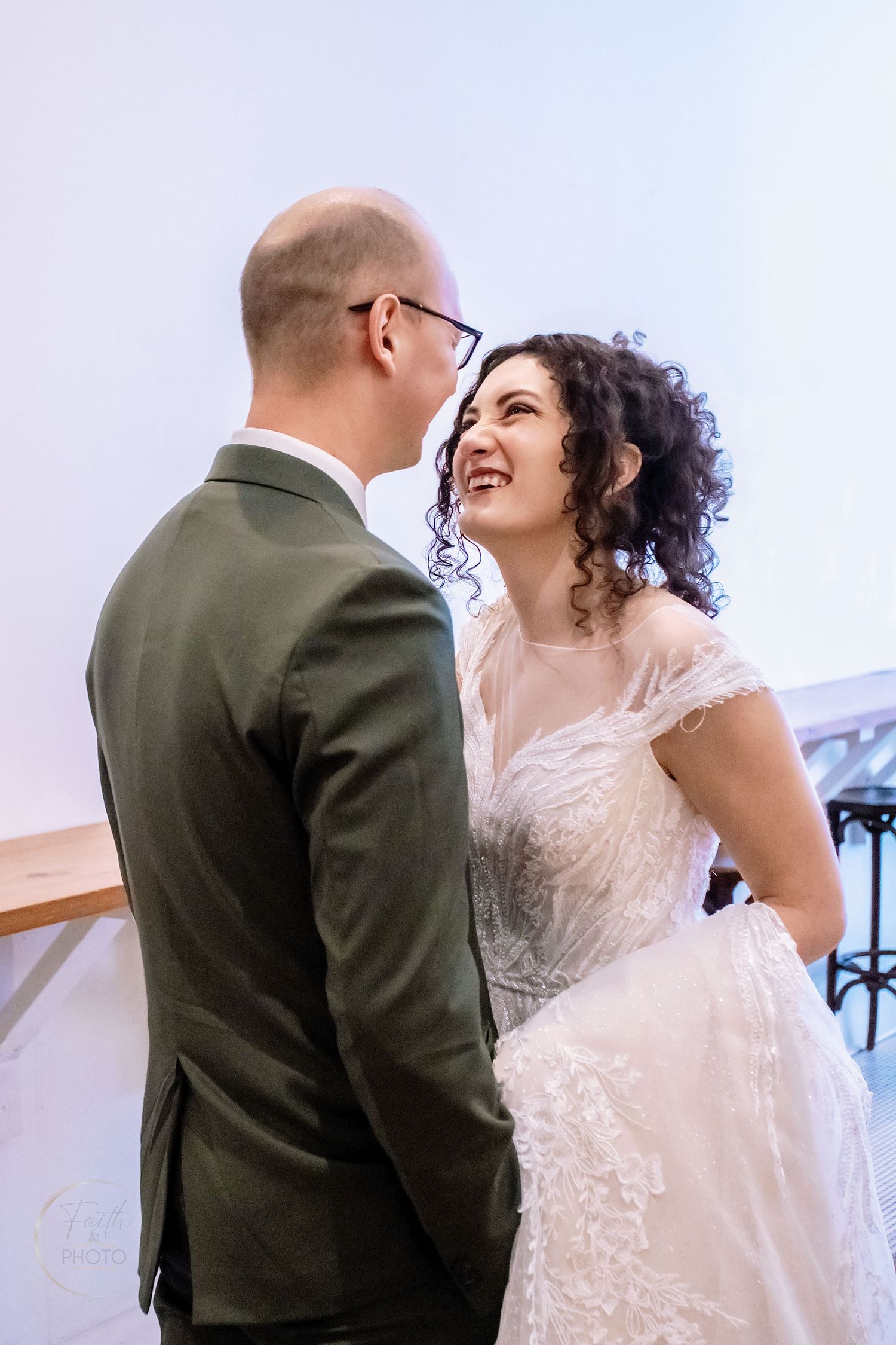 Smiling bride in lace gown gazes at bald groom in green suit during wedding ceremony indoors.