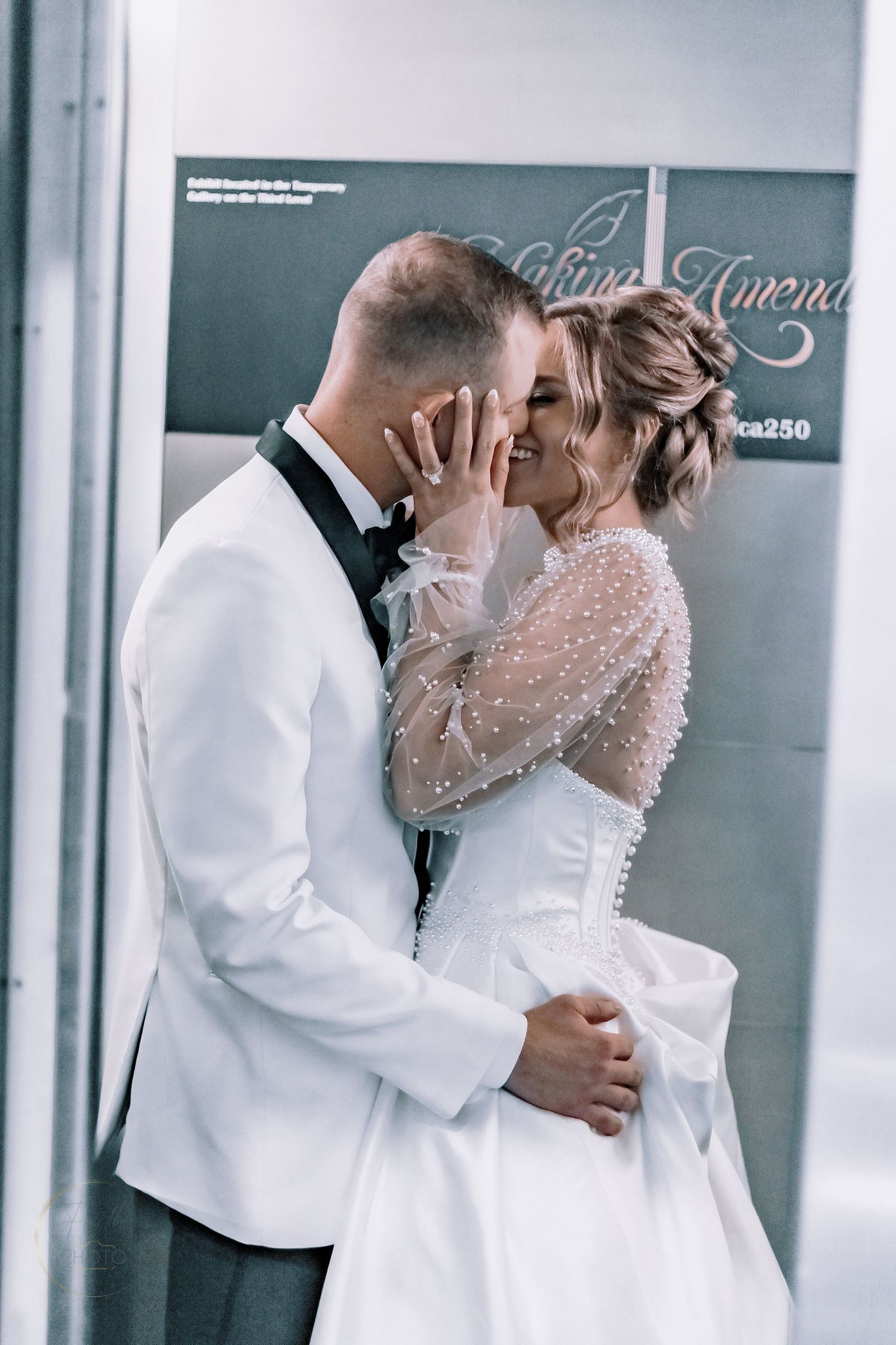 Bride and groom share a romantic kiss in wedding attire, she in a pearl-adorned gown, he in a white tuxedo jacket.