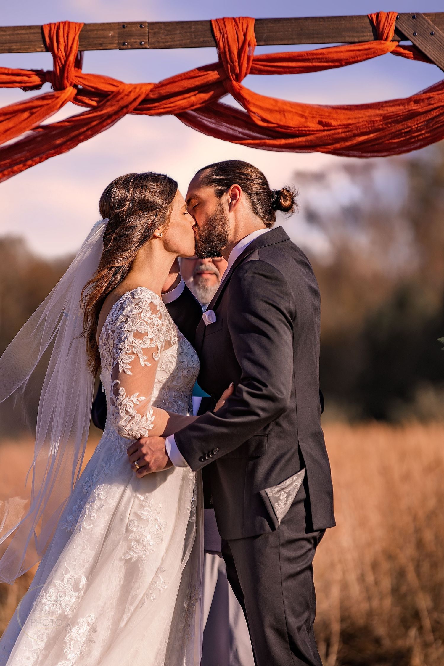 Bride and groom share first kiss under red draped arch at outdoor wedding ceremony.