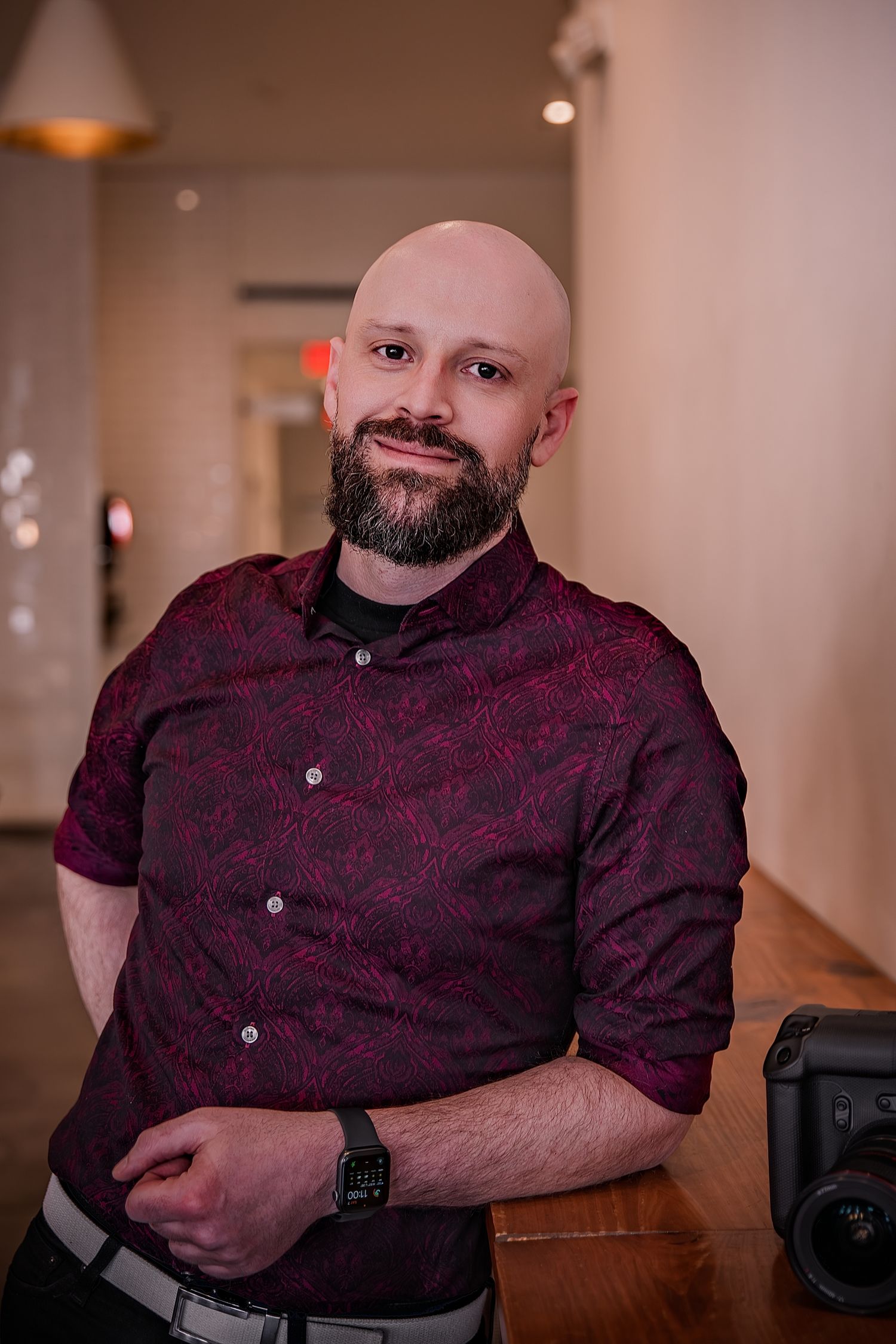 Bald man with beard wearing dark red patterned shirt, smiling with arms crossed, standing near wooden table indoors.
