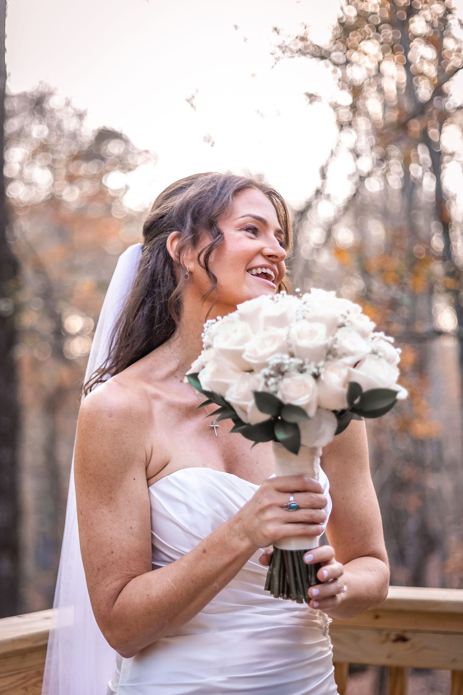 Joyful bride in white dress laughs while holding a bouquet of white roses in an autumn outdoor setting.