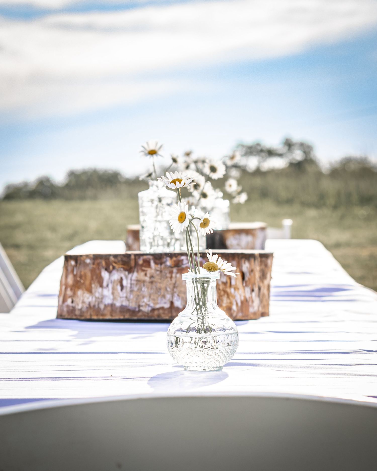 White daisies in a glass vase sit on a rustic wooden table with a white tablecloth at an outdoor event setting.