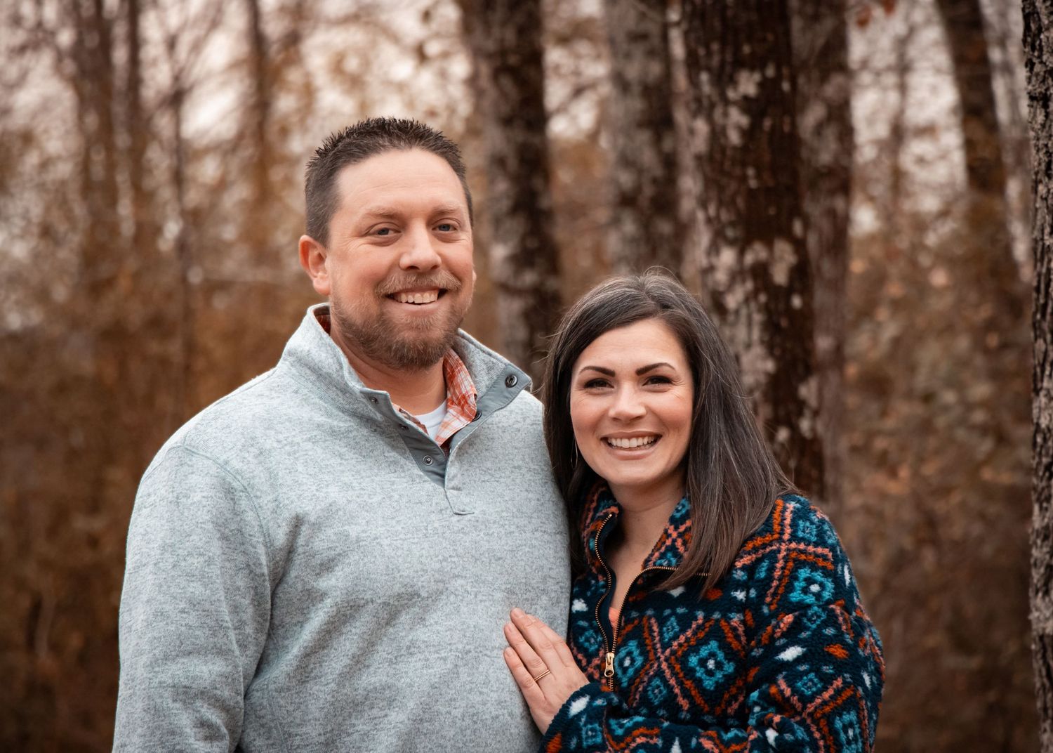 A couple stands close together smiling outdoors in an autumn forest setting.