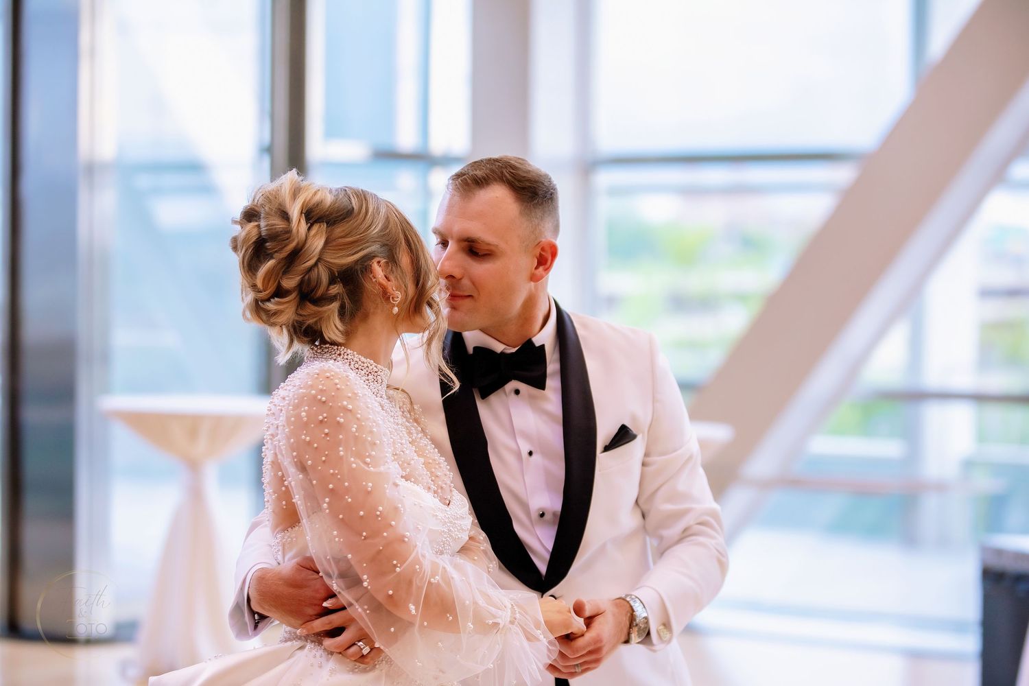 Bride and groom share a romantic moment in a bright venue, groom in white tuxedo, bride in lace wedding gown.
