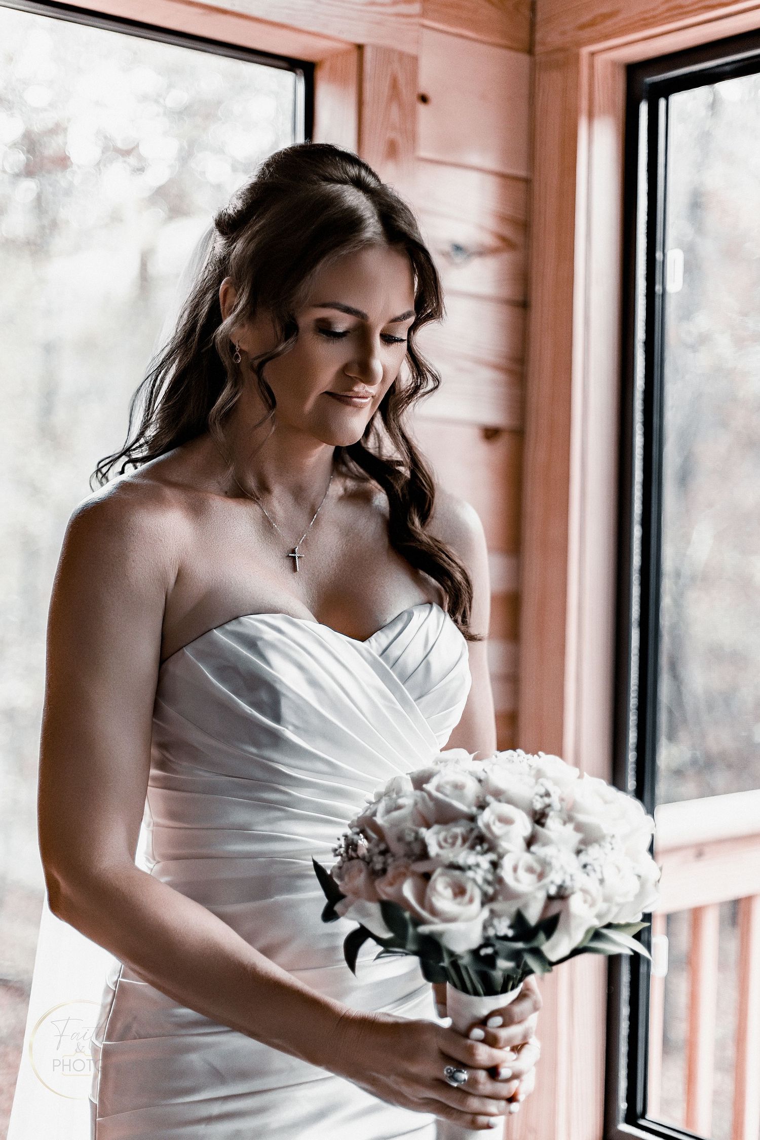 Bride in strapless white gown holds white rose bouquet by window in wooden venue, looking down gracefully.