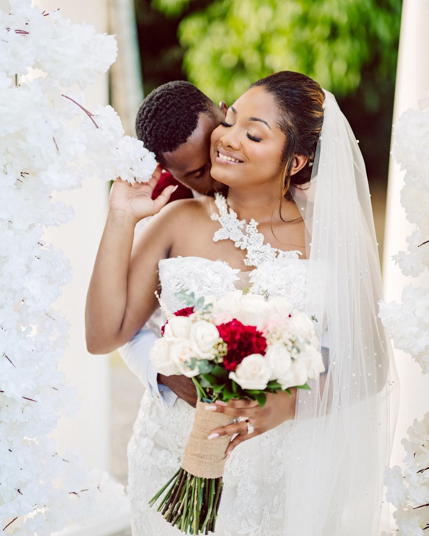 Bride holding red and white bouquet while sharing intimate moment during wedding photoshoot.