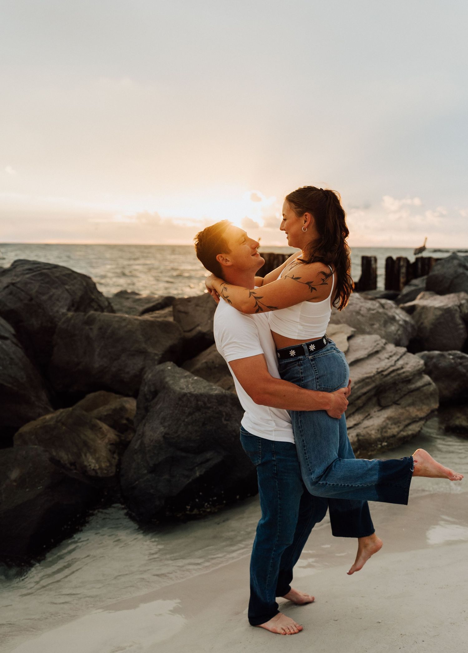 A romantic couple embraces on a rocky beach at sunset creating a silhouette against the ocean backdrop.