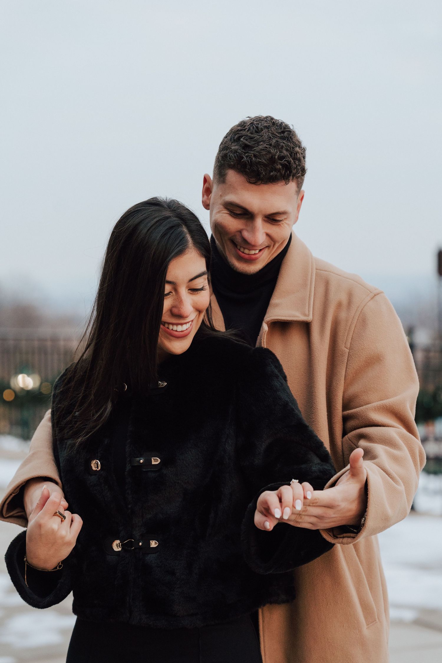 A couple laughs and embraces while looking at the engagement ring during a winter engagement photoshoot.