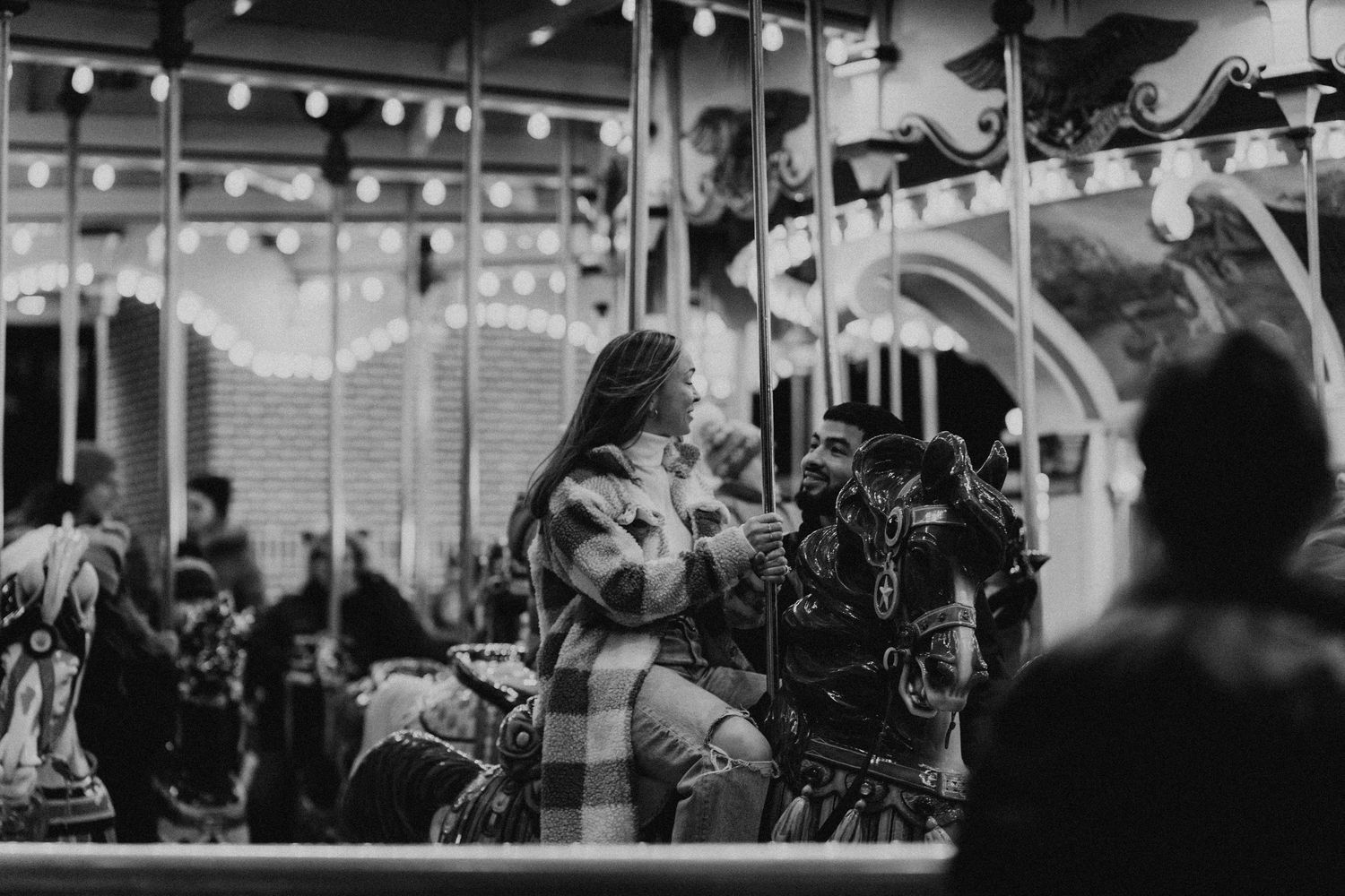 Black and white photo of engaged couple smiling at each other while riding the carousel in Hershey Park for engagement photoshoot.