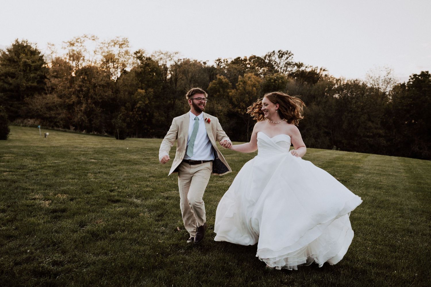 Newlyweds running joyfully through a grassy field at sunset.