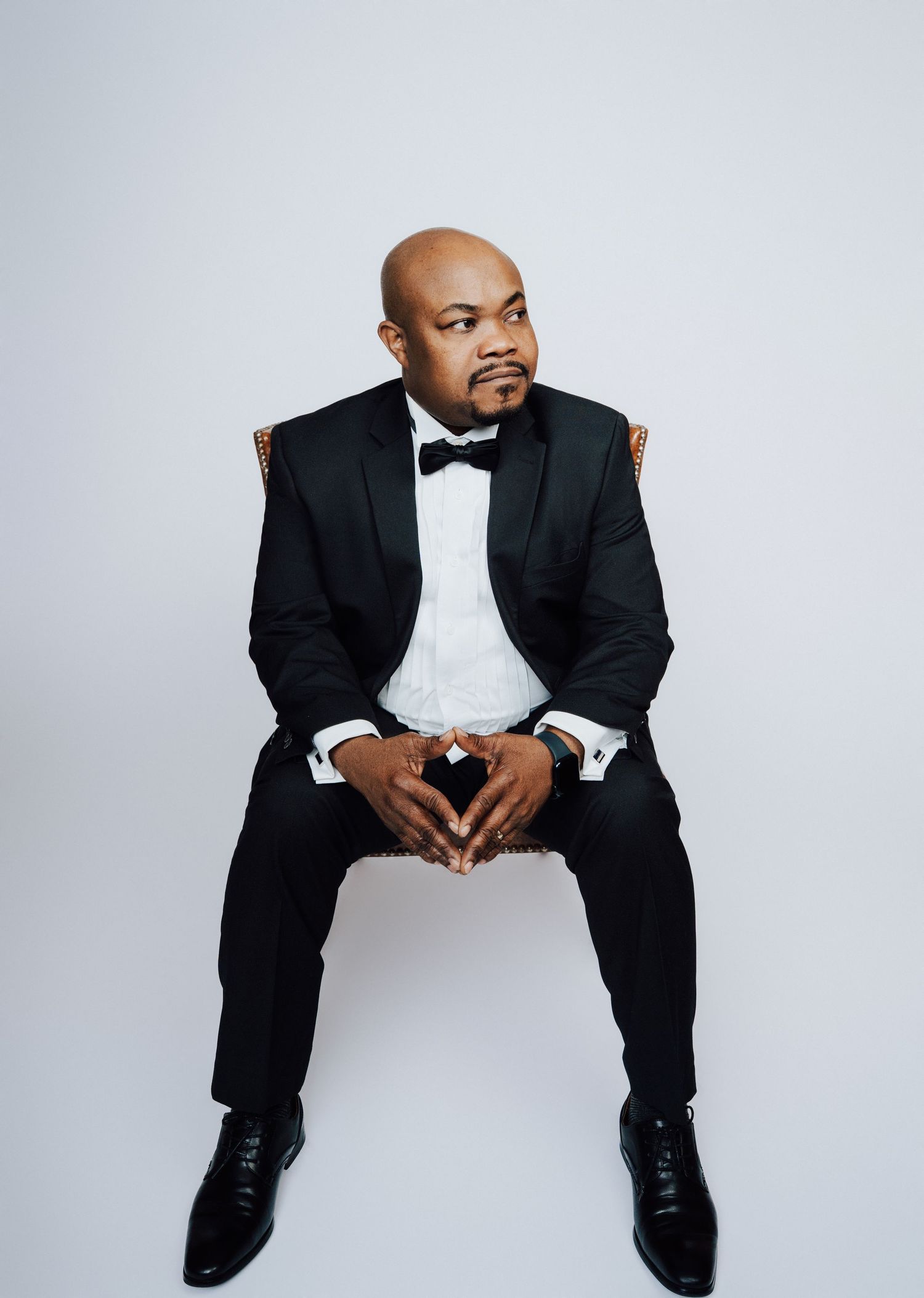Bald man in black tuxedo with bow tie sits confidently against white background.