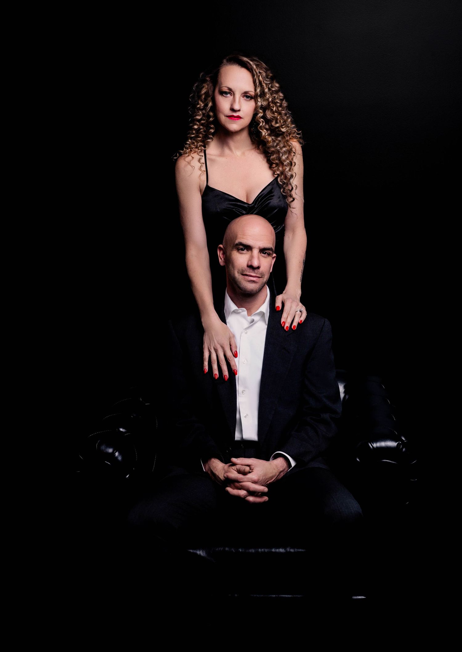A woman in a black dress stands behind a seated bald man in a suit against a dark background.
