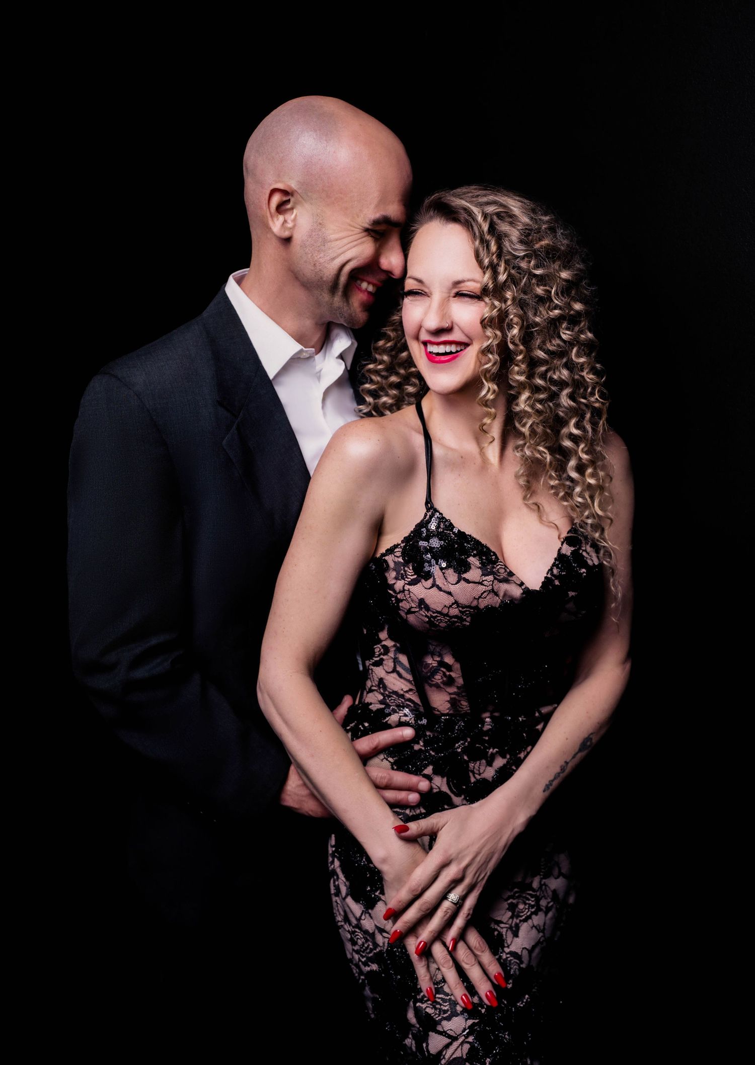 Elegant couple posing together against black background, bald man in suit embracing smiling woman in black lace dress.