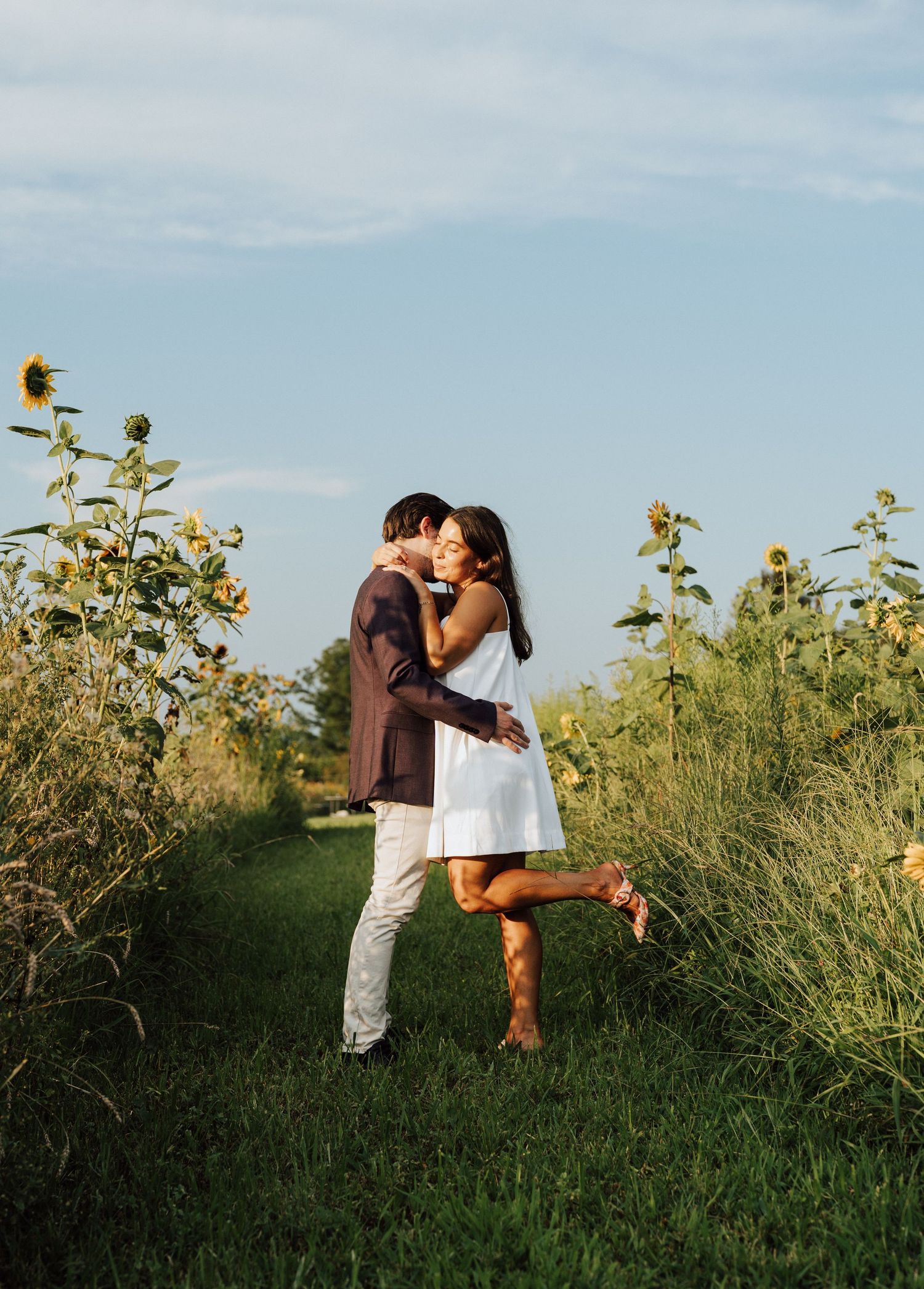 A romantic couple embraces in a sunflower field on a summer day, with the person in white lifting one foot.