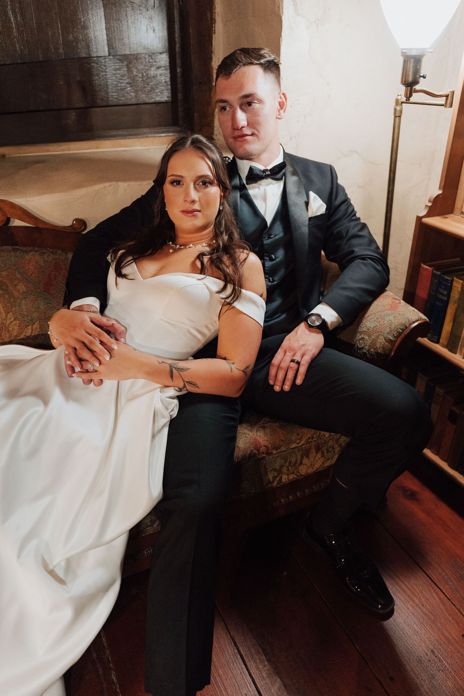 Newlywed couple sits together on vintage couch, bride in white off-shoulder gown and groom in classic black tuxedo.