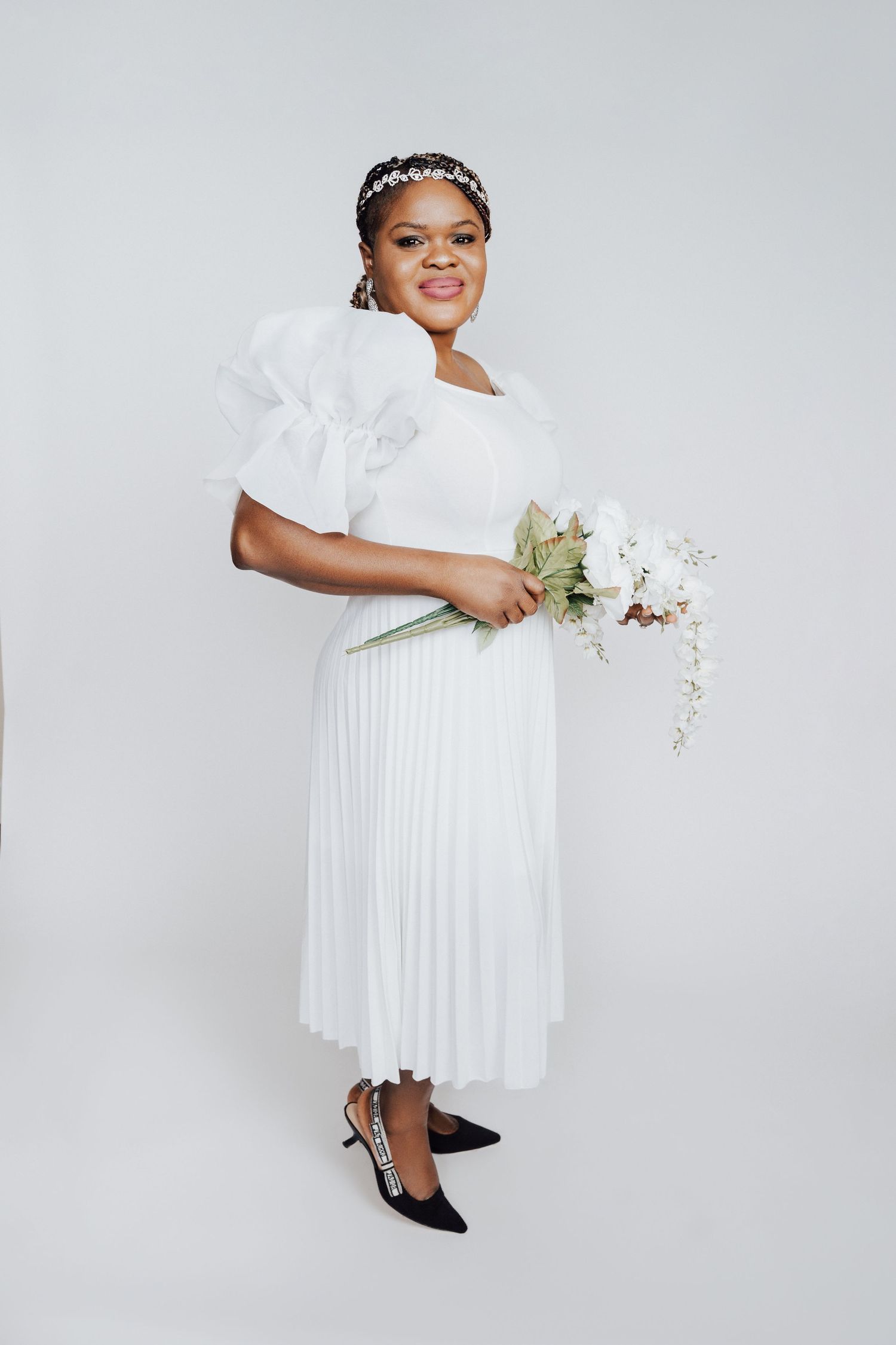 Black bride in white pleated midi dress with puff sleeves, floral headband, holding white bouquet, posing on white background.
