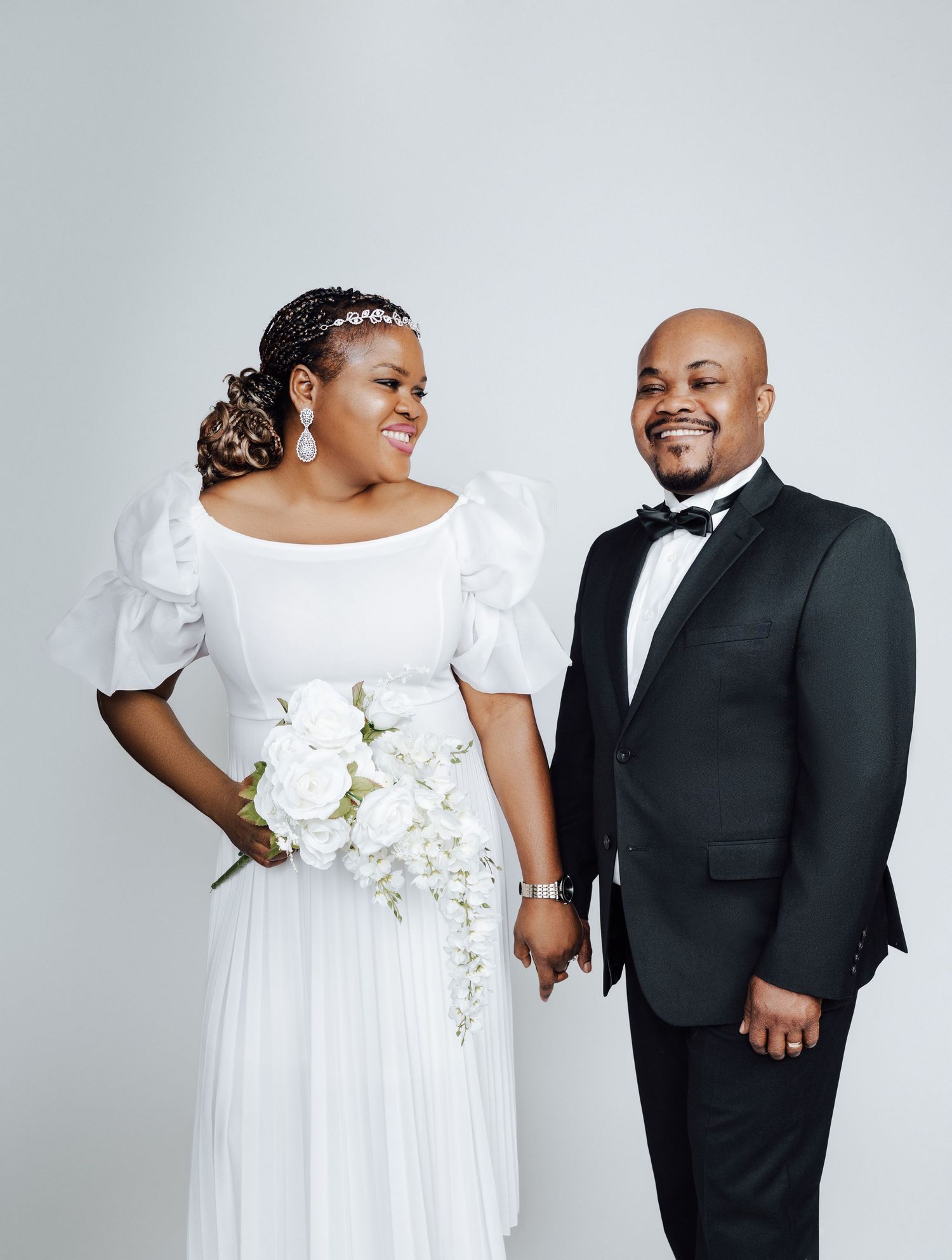 A bride in white gown holding bouquet and groom in black tuxedo pose together smiling at their wedding.