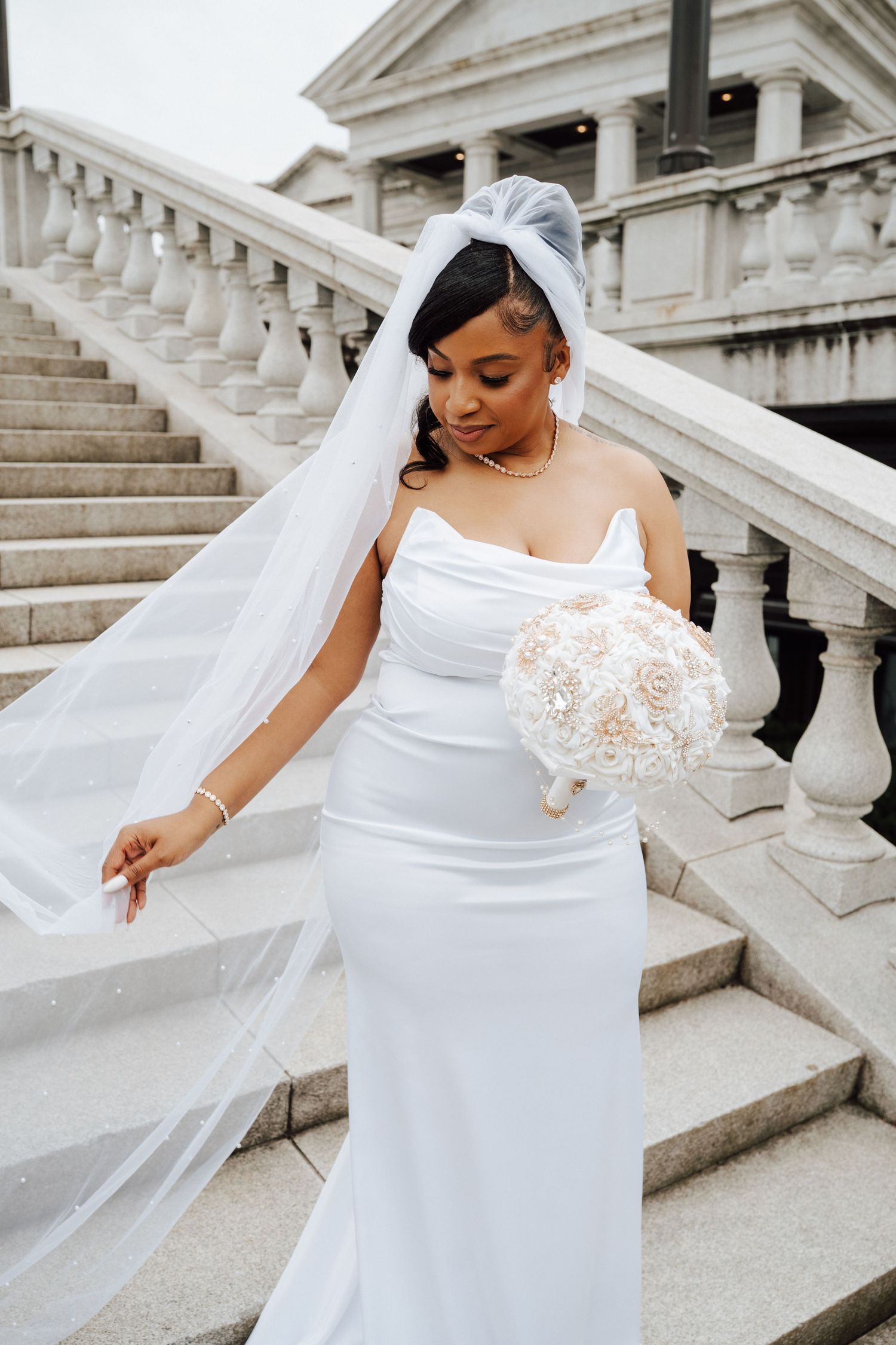 Bride in elegant white gown with flowing veil poses gracefully on grand marble staircase in wedding photo series.