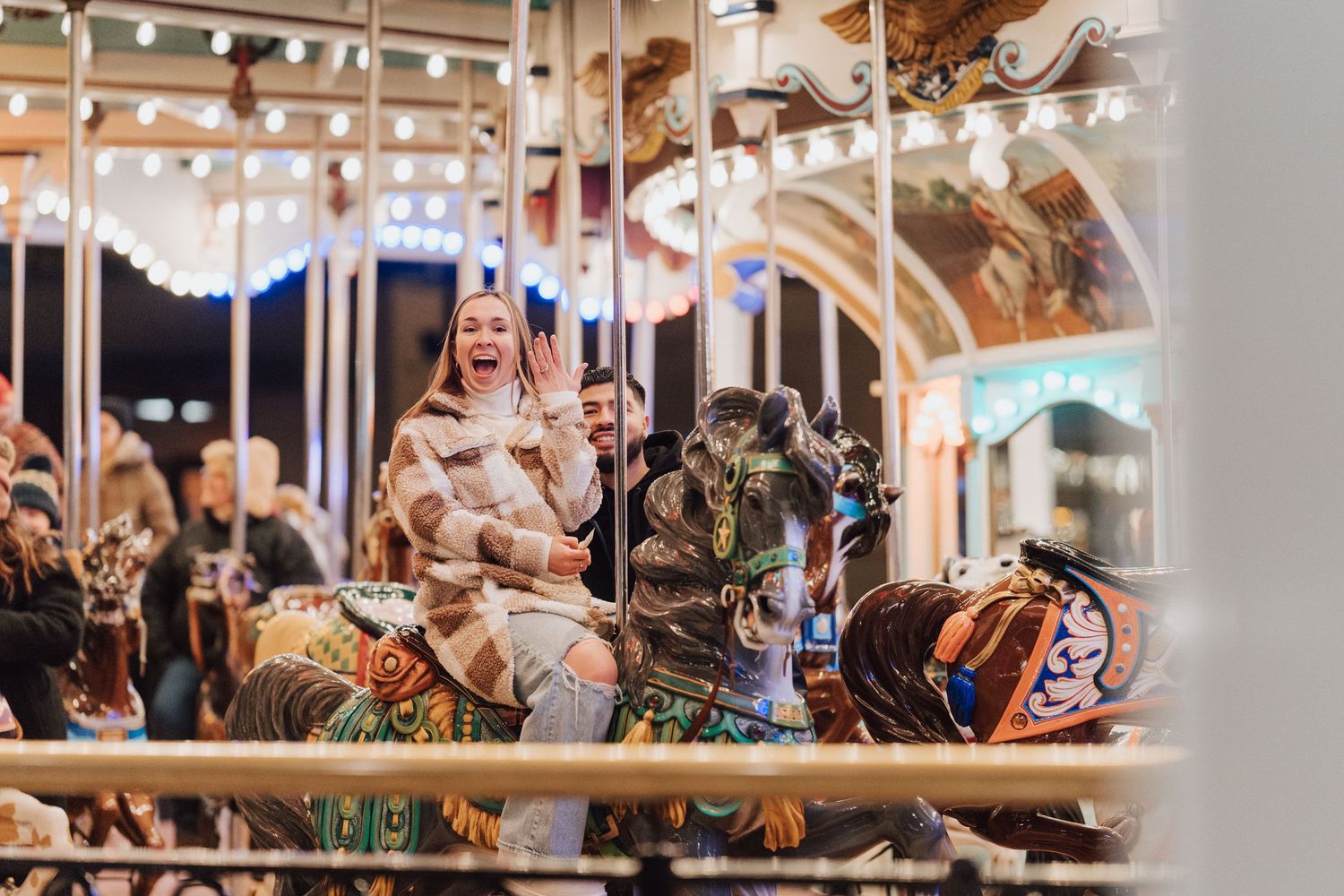Woman showing with excitement her engagement ring while riding the carousel in Hershey Park, Hershey, PA for engagement proposal photoshoot.