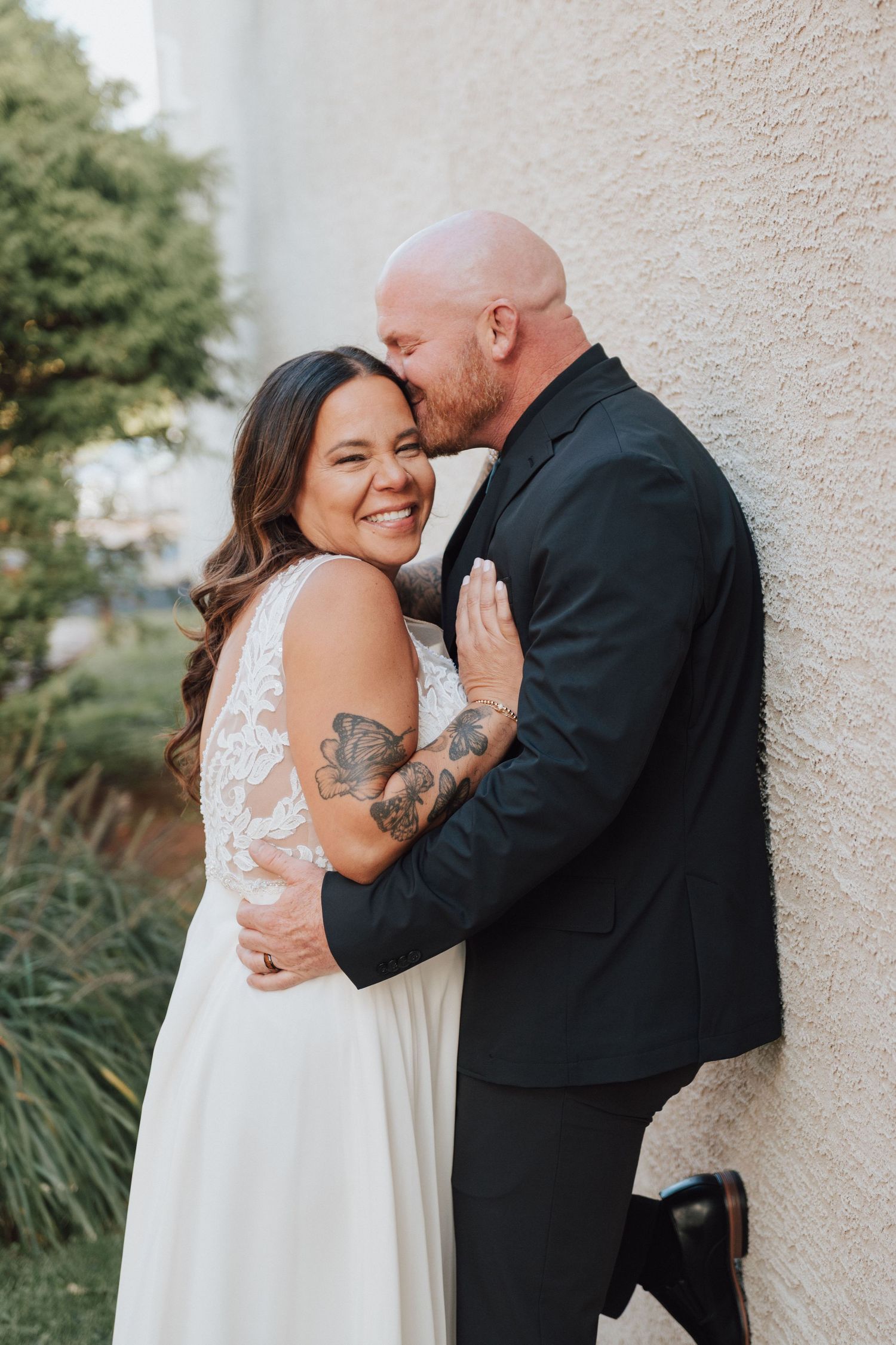 A hispanic bride with tattoos in one arm smiling as her husband is kissing her forehead on their wedding day.
