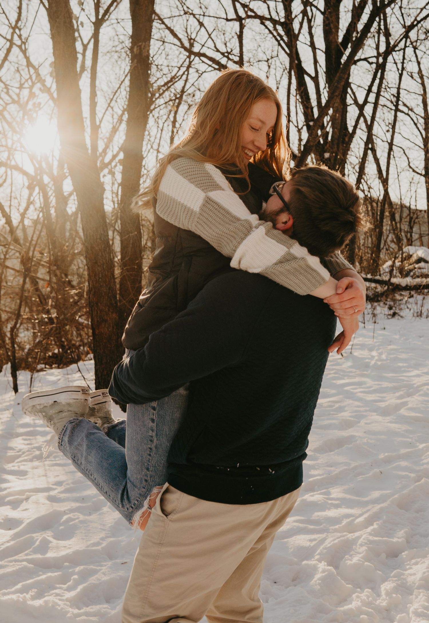Two people embracing in a snowy winter forest with golden sunlight streaming through bare trees.