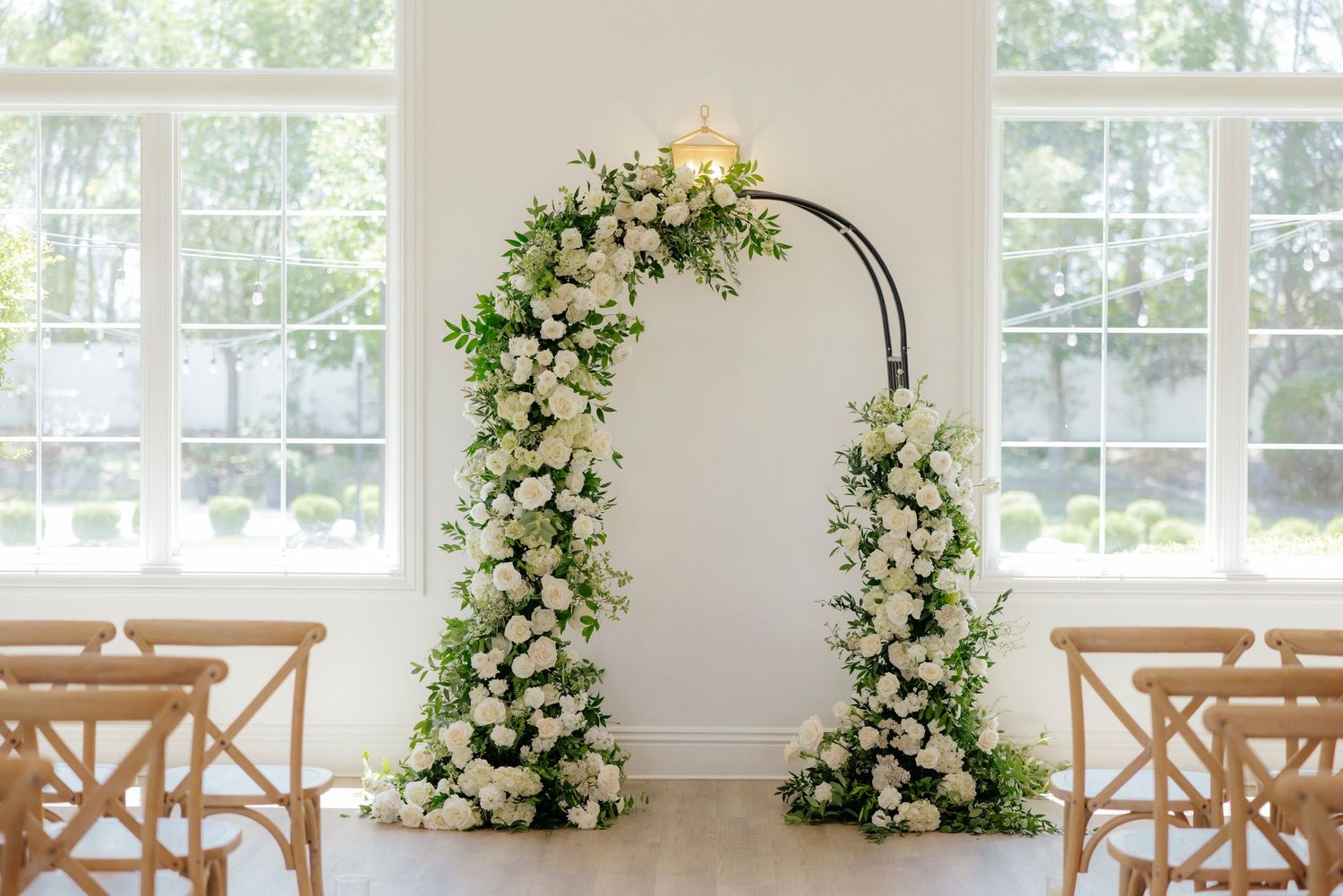 Elegant white wedding chapel interior with wooden cross-back chairs and floral arch arrangements by large windows.