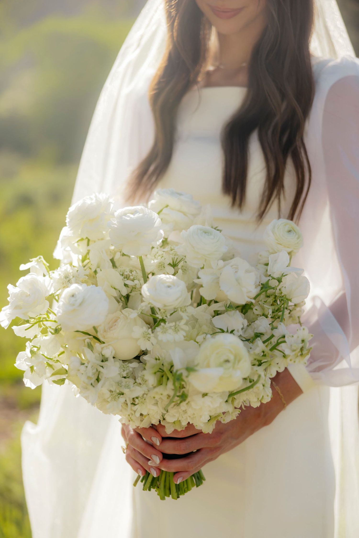Bride in white gown and veil holding a lush bouquet of white ranunculus and hydrangeas outdoors.