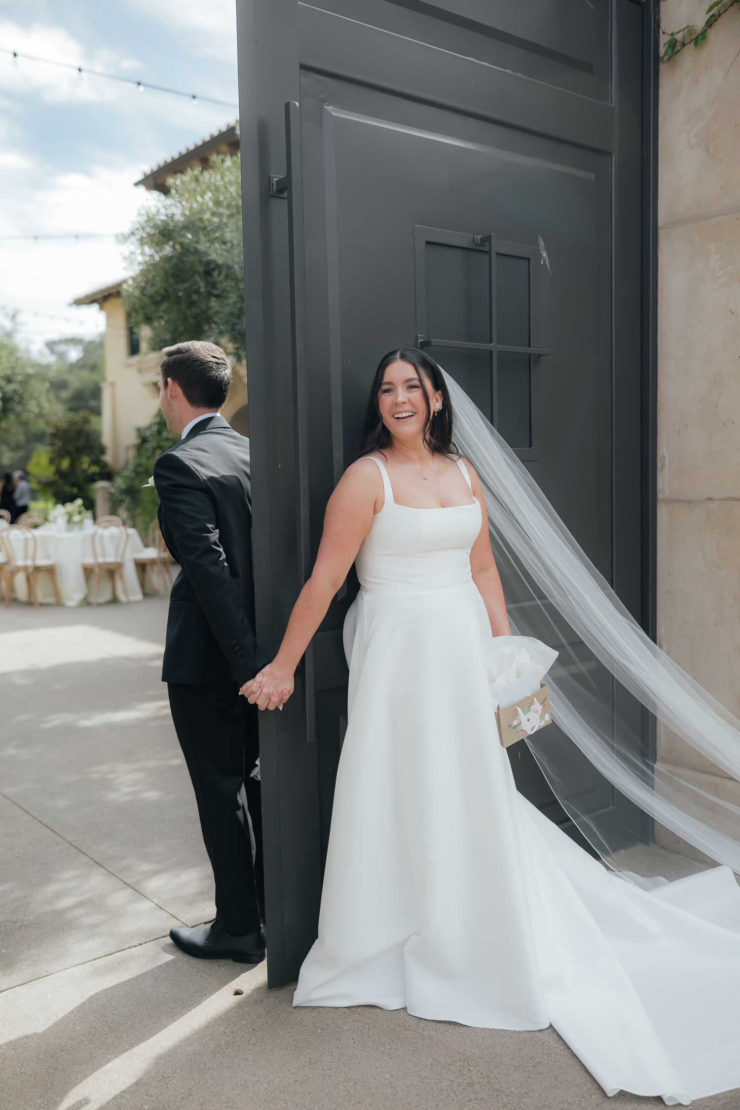 Bride in white gown and veil holds groom's hand outside elegant venue doors on their wedding day.