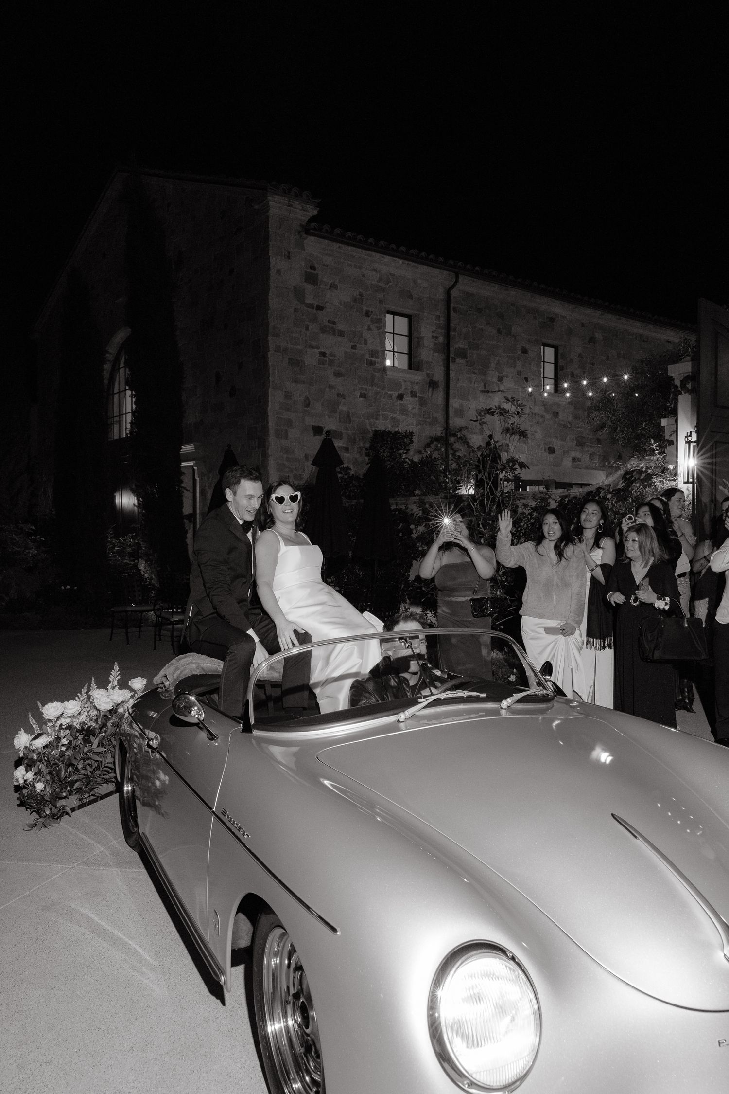 Bride and groom exit wedding in vintage Porsche convertible at night, guests cheering, stone venue backdrop.