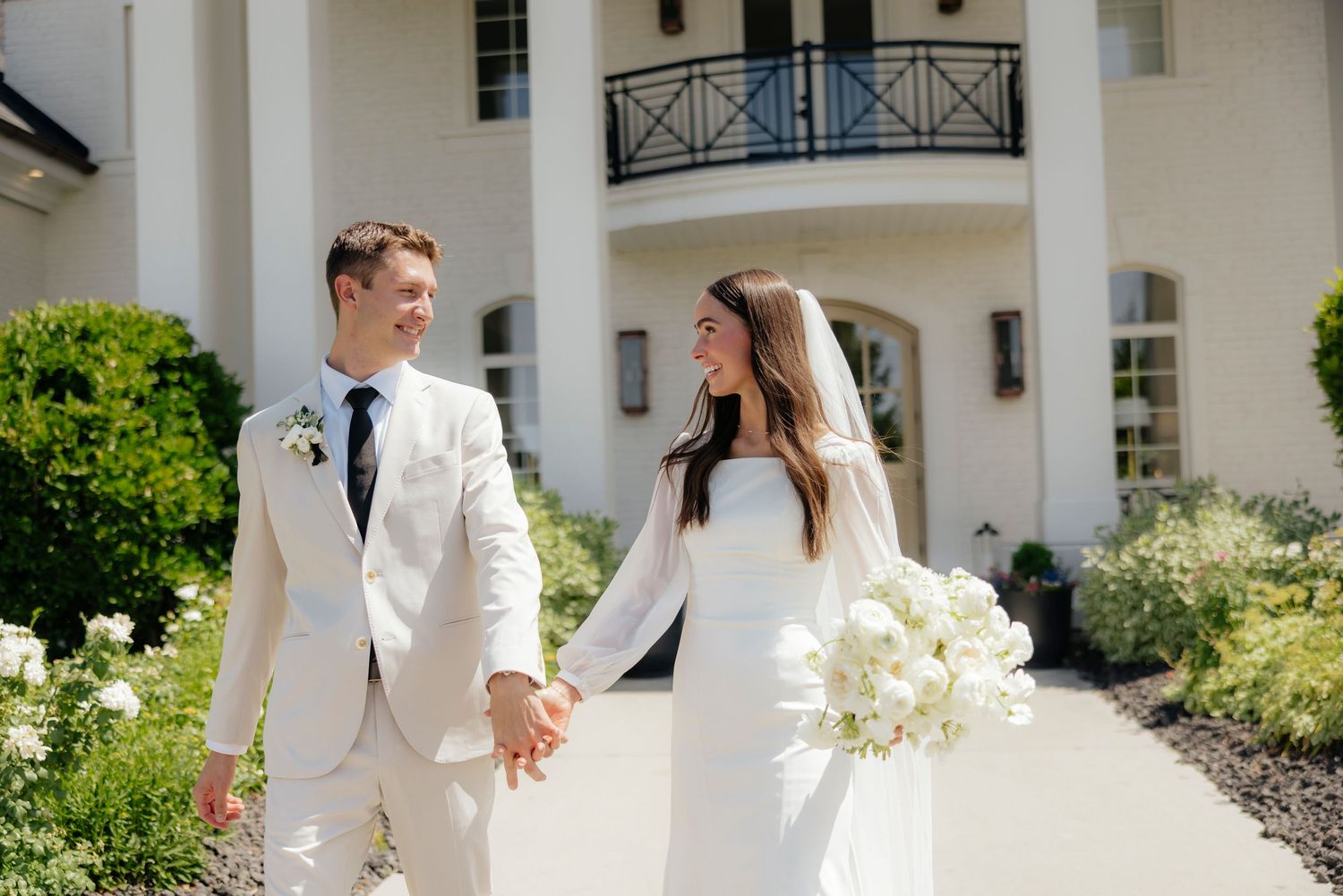 A couple in elegant white attire walking together through a garden path on a sunny day.