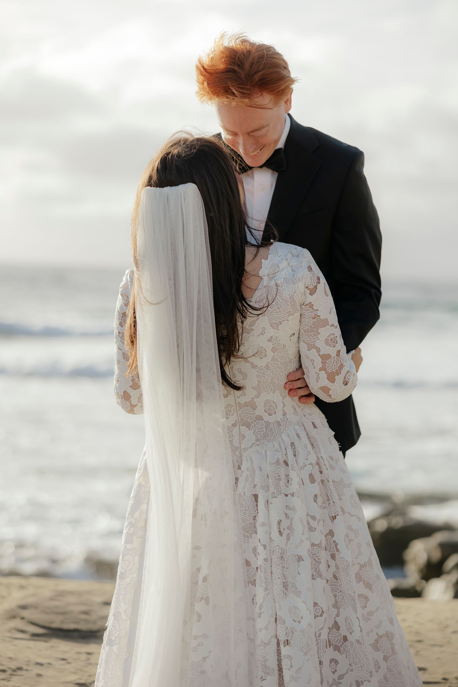 A couple in wedding attire share an intimate moment on a rocky beach at sunset with waves in the background.