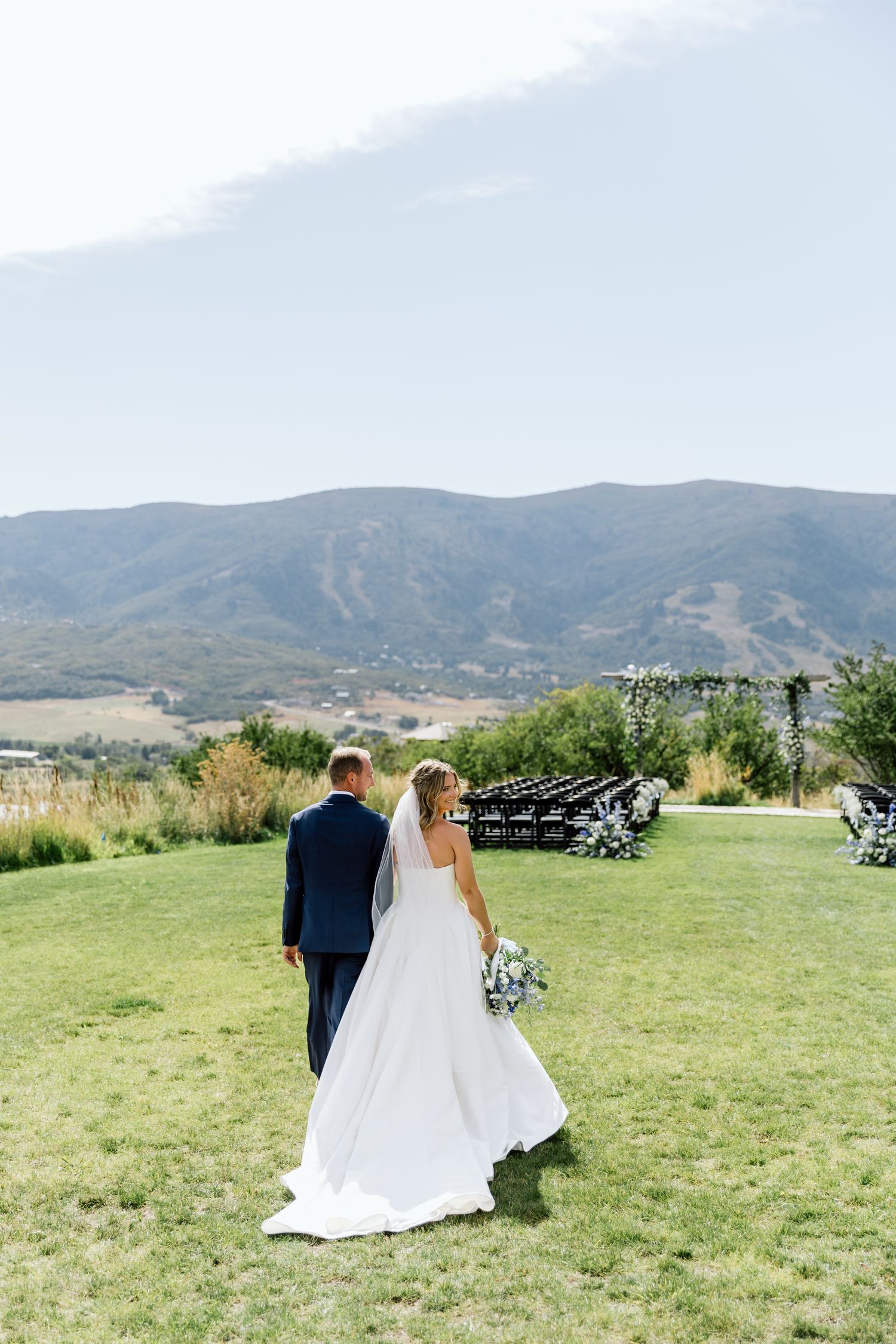 A couple in wedding attire walks across a scenic mountain meadow with rolling hills in the background.