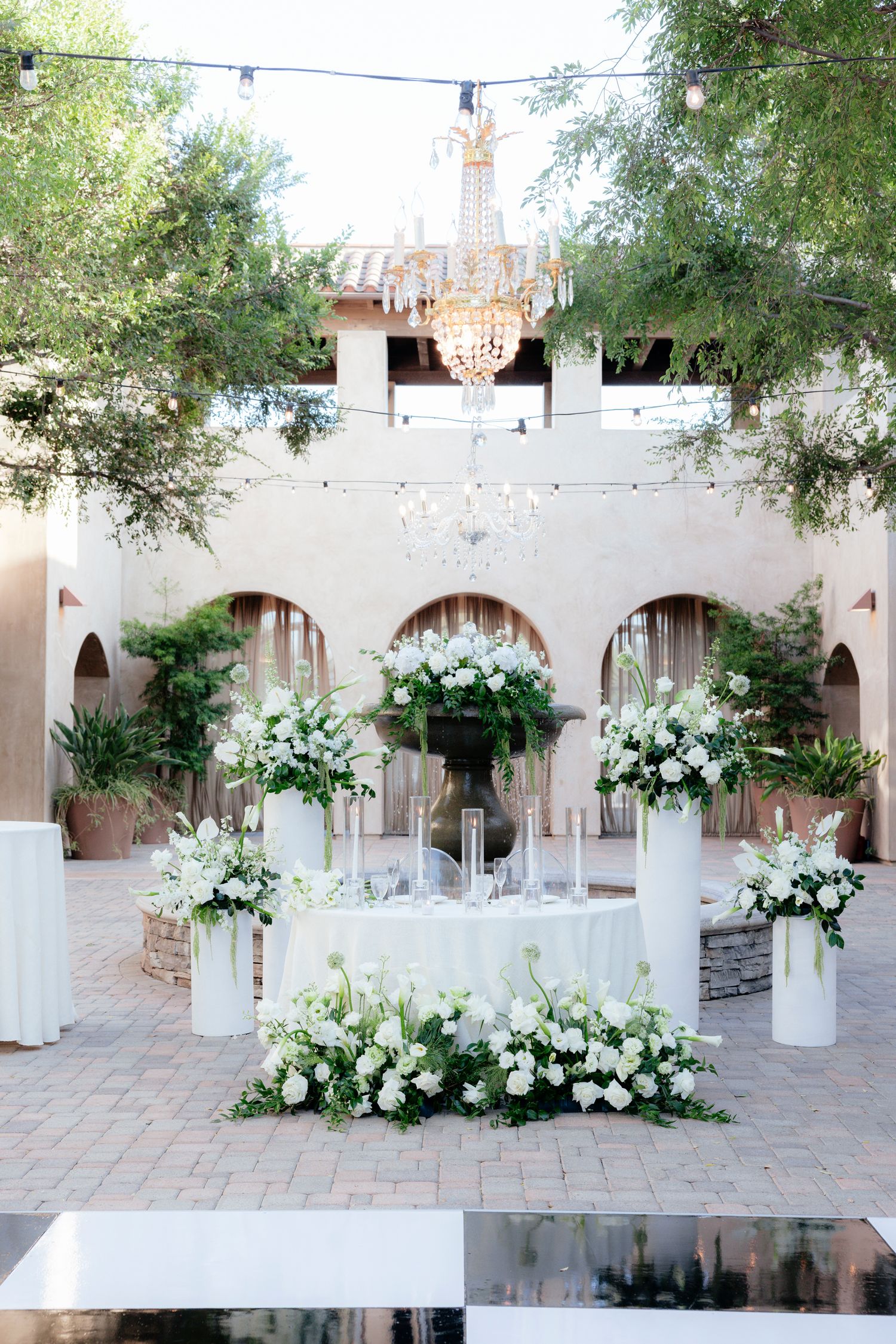 Elegant outdoor wedding reception with white floral arrangements, chandelier, and string lights in a courtyard venue.