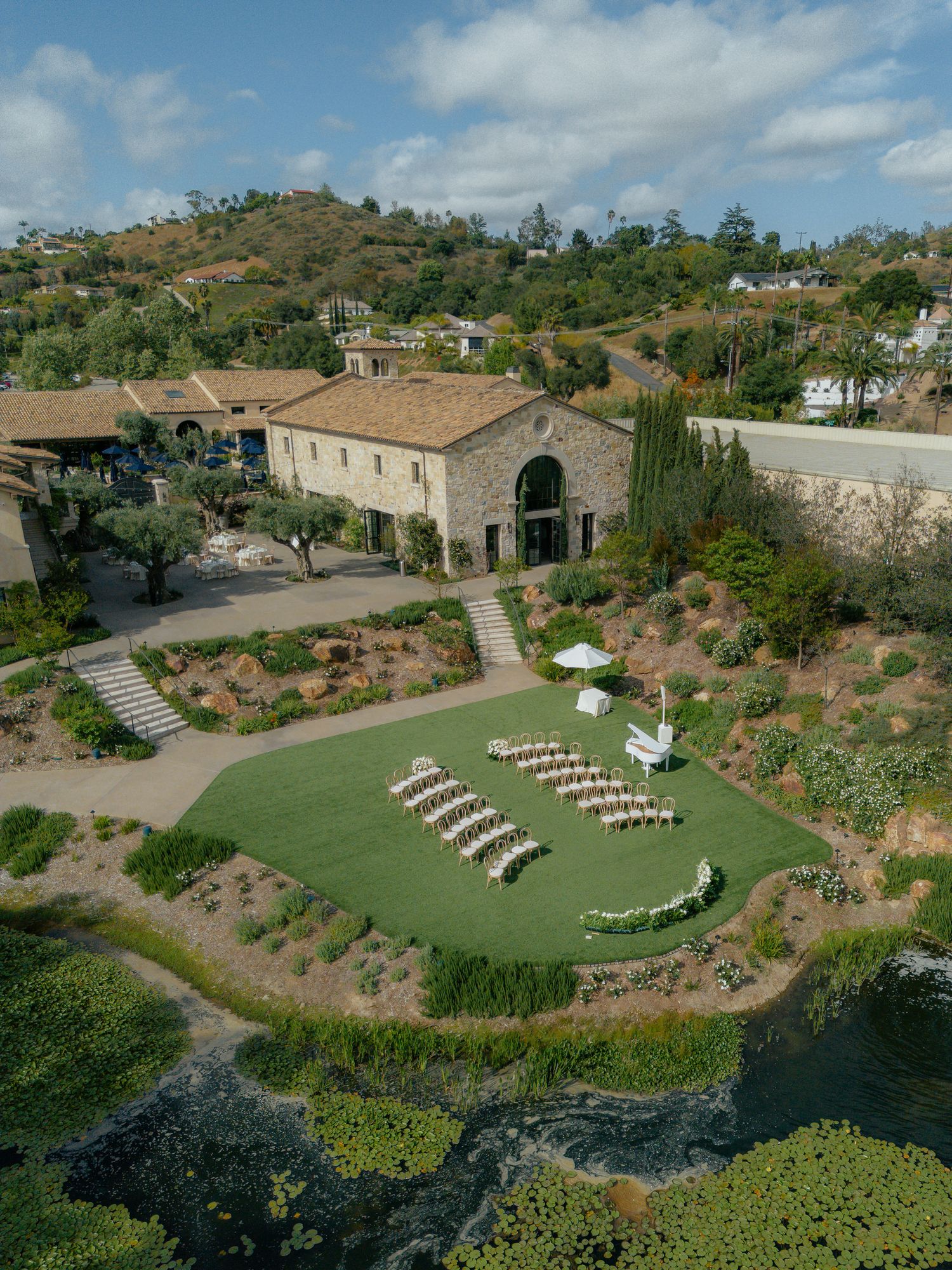 Aerial view of an elegant outdoor wedding ceremony on a green lawn beside a Tuscan-style villa surrounded by lush hills.