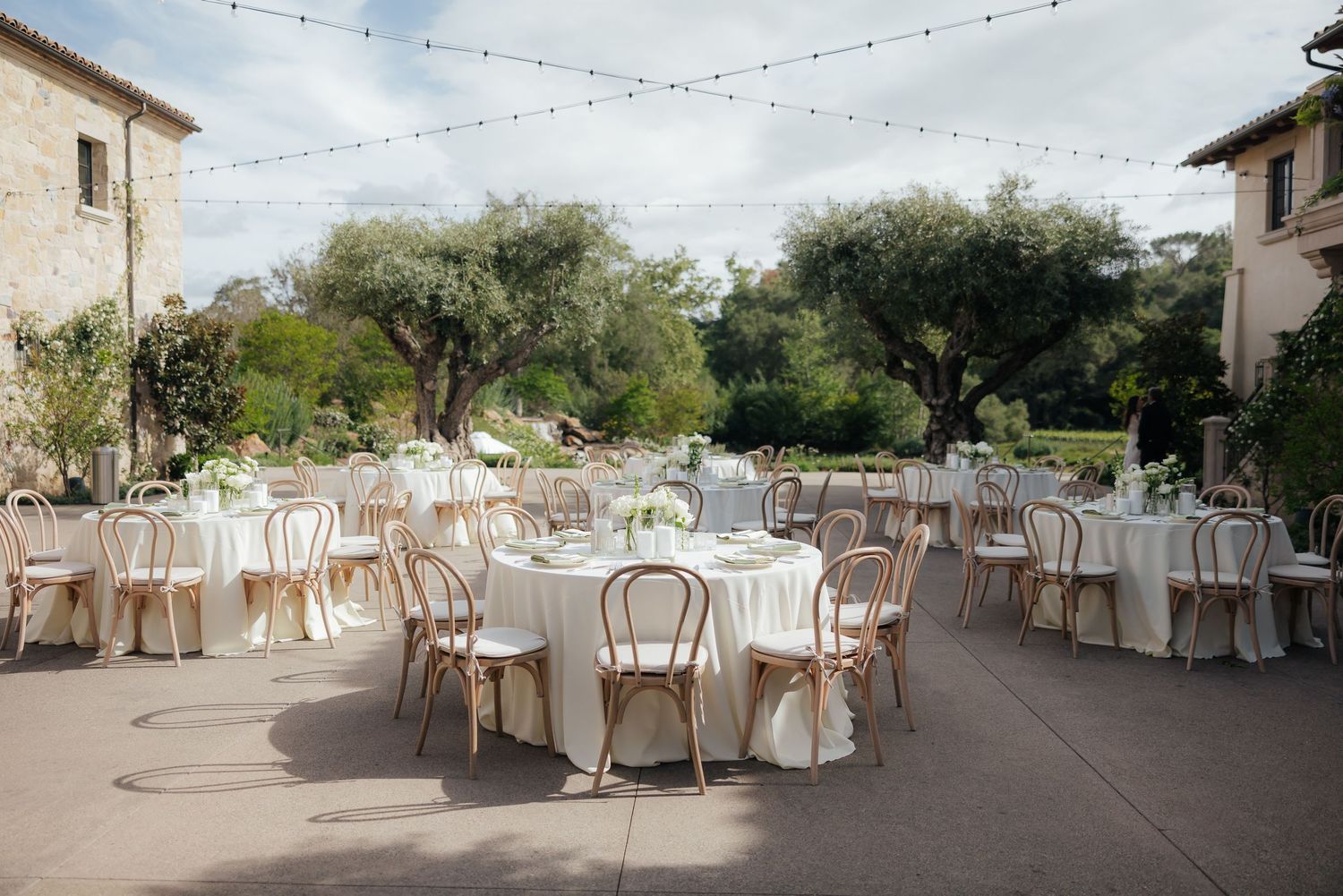 Elegant outdoor wedding reception with round tables, white linens, wooden chairs, string lights, and lush greenery at a rustic venue.