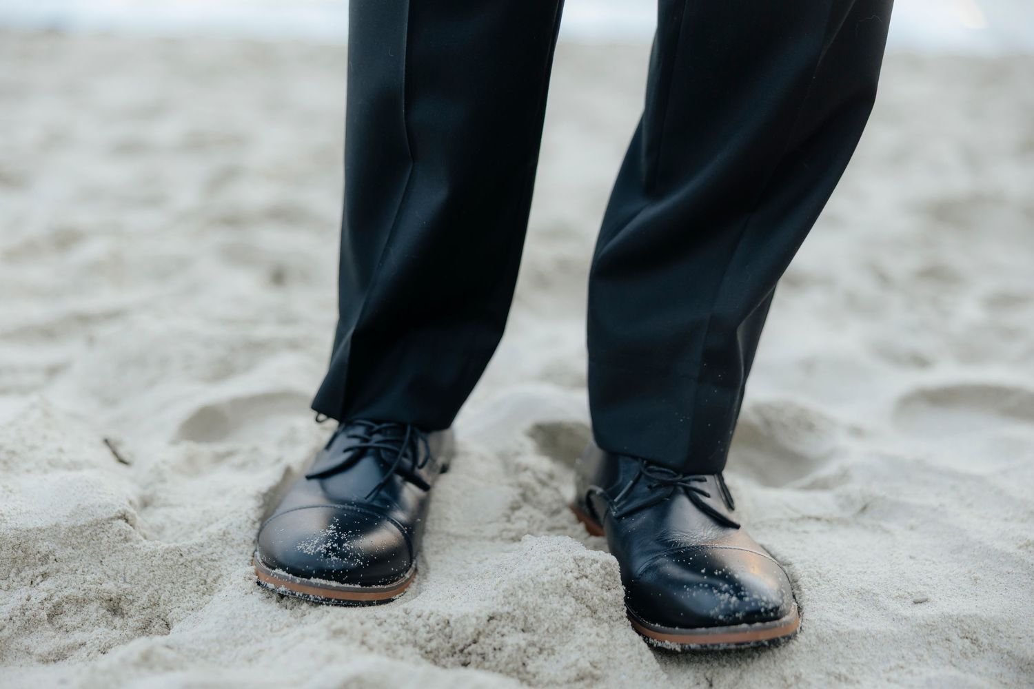 Black dress shoes and suit pants standing in soft beach sand with footprints visible.