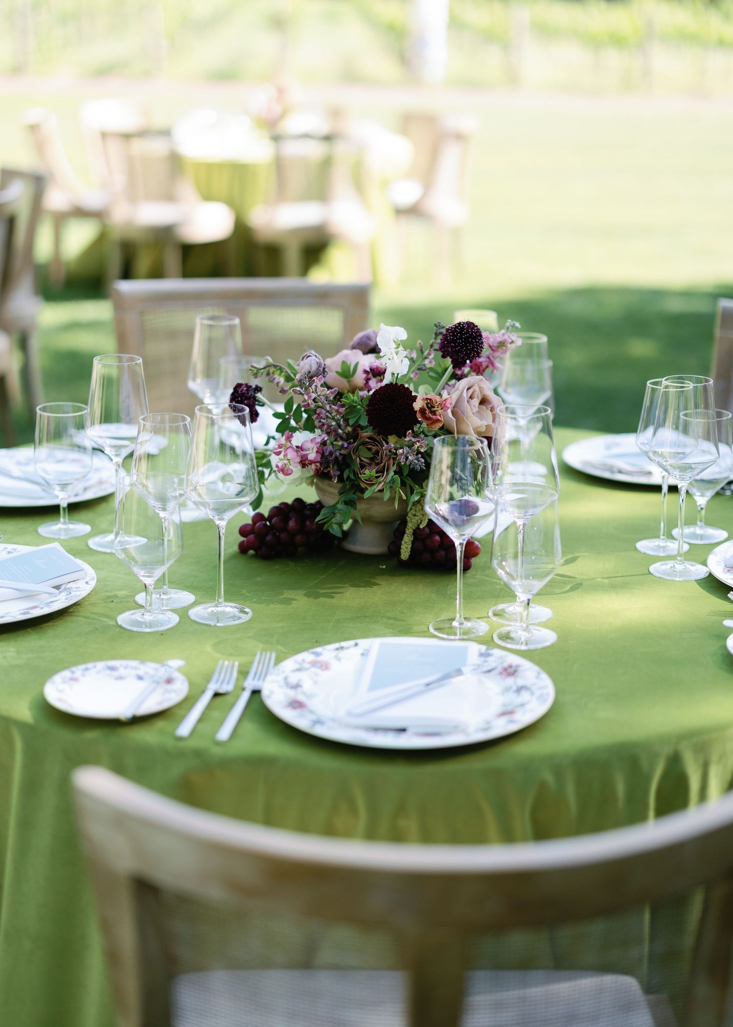 Elegant outdoor wedding table with green linen, crystal glasses, floral centerpiece of burgundy and blush blooms.