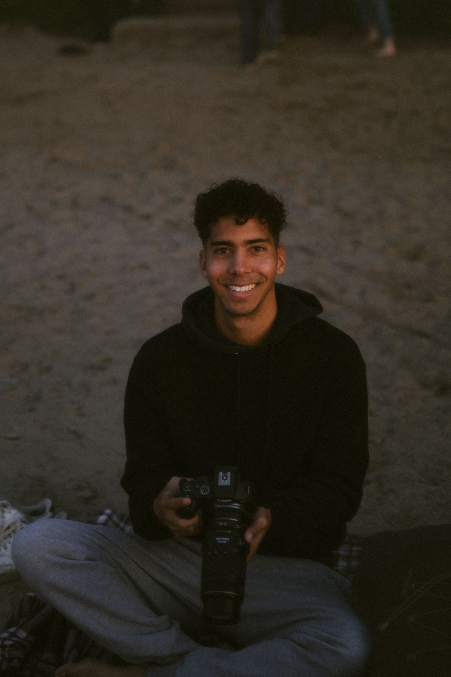 Smiling young man sitting on beach sand holding DSLR camera, wearing black hoodie and grey pants.