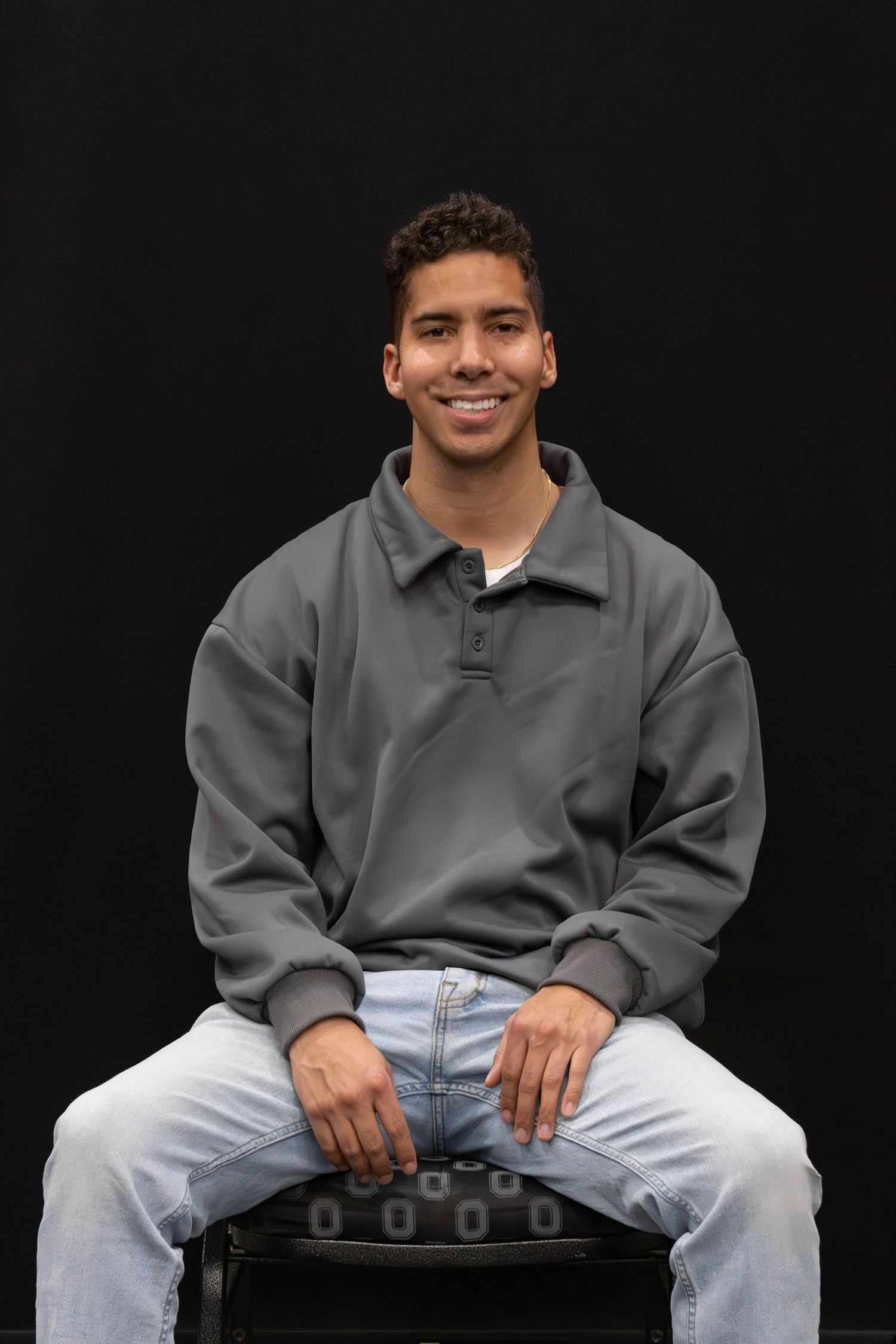 Young man in gray sweatshirt and jeans sitting on chair smiling against a black studio background.