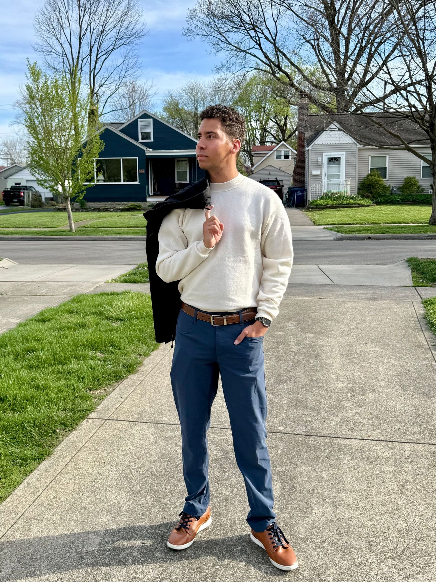 A young man in a cream sweater and blue pants poses on a suburban sidewalk on a clear spring day.