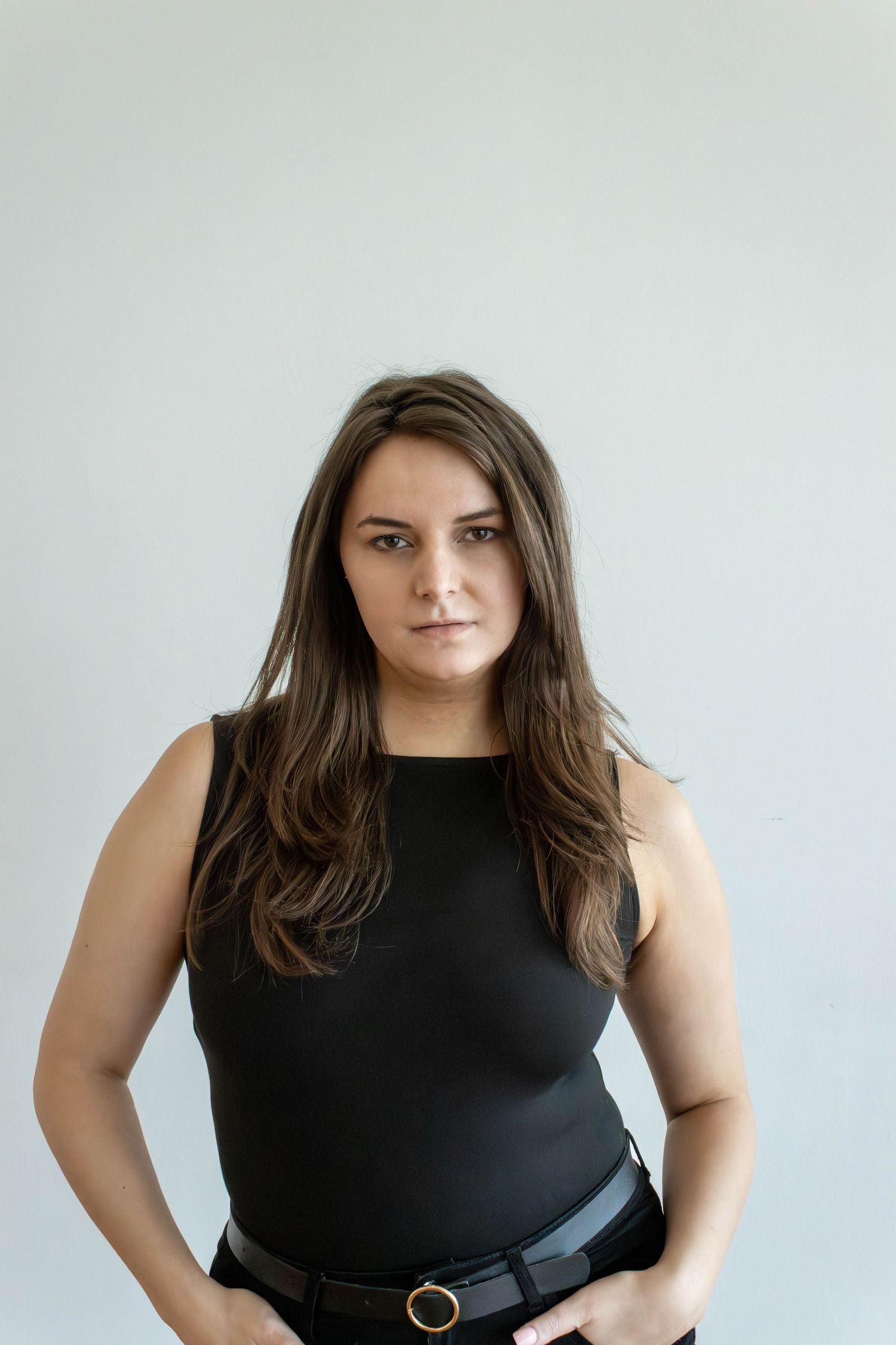 Young woman with long brown hair wearing a black sleeveless top and belt, posing confidently against a white background.