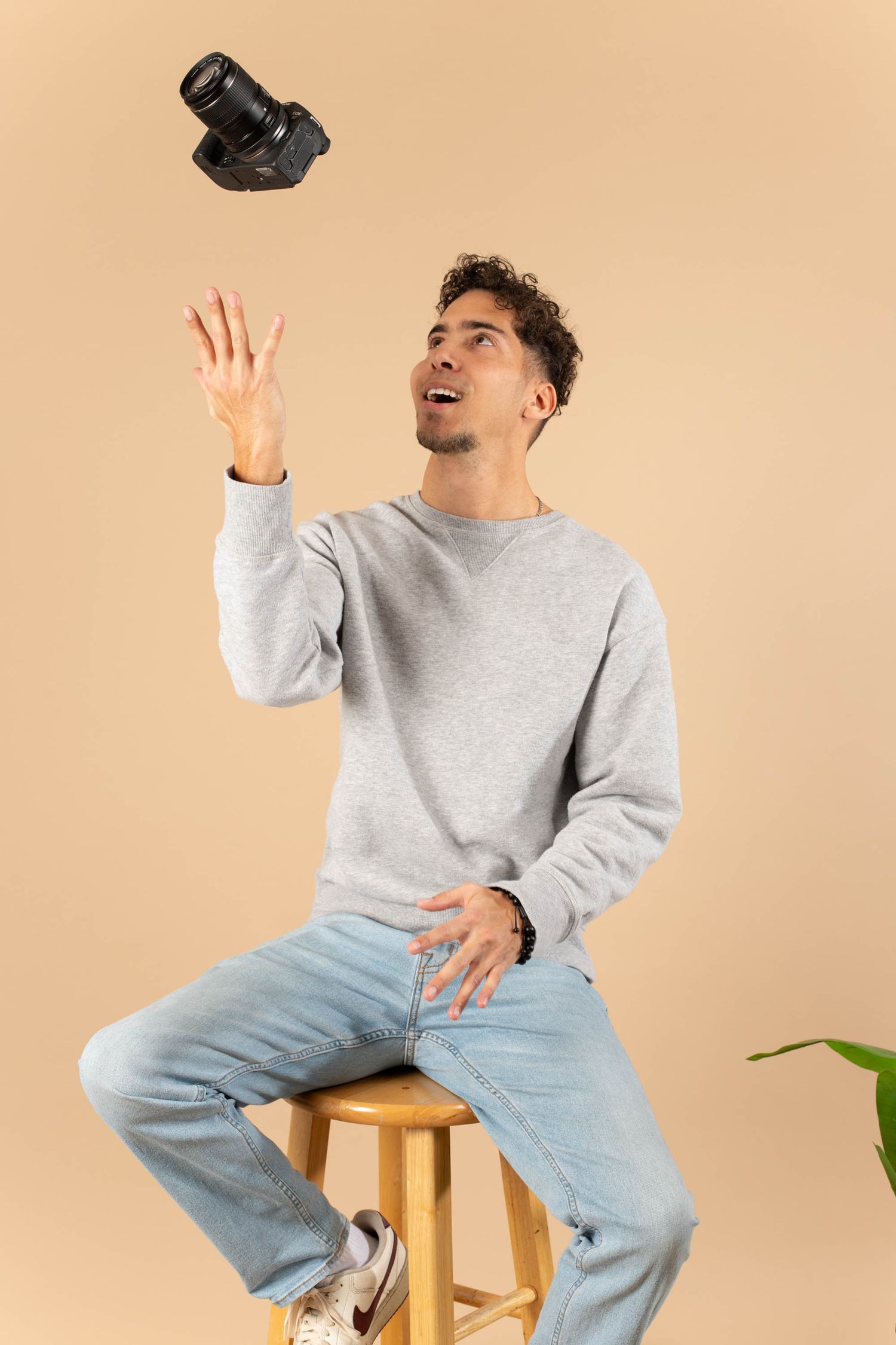 Young man sitting on stool tossing DSLR camera in air, wearing grey sweatshirt and jeans against beige background.