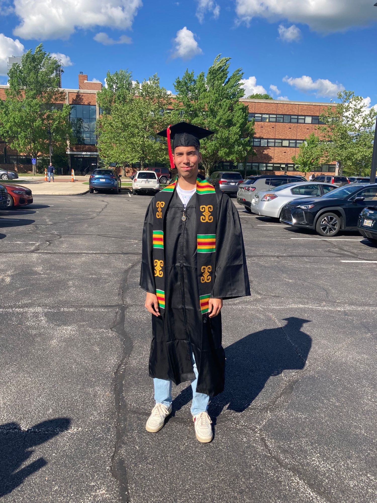 Graduate in black cap and gown with kente cloth stole standing in sunny parking lot.
