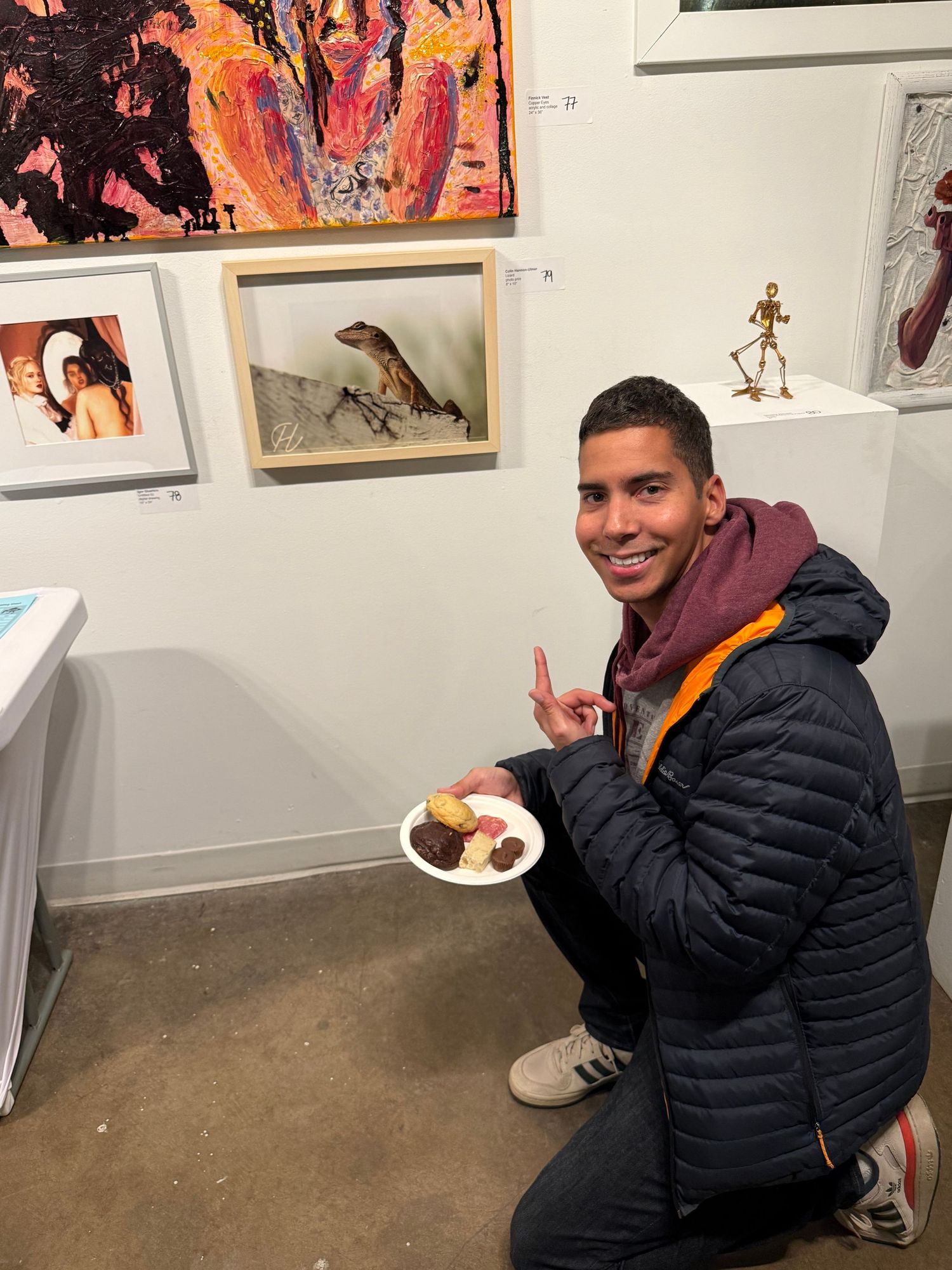 Young man smiling and holding a plate of cookies while crouching in front of colorful artwork at an art gallery.