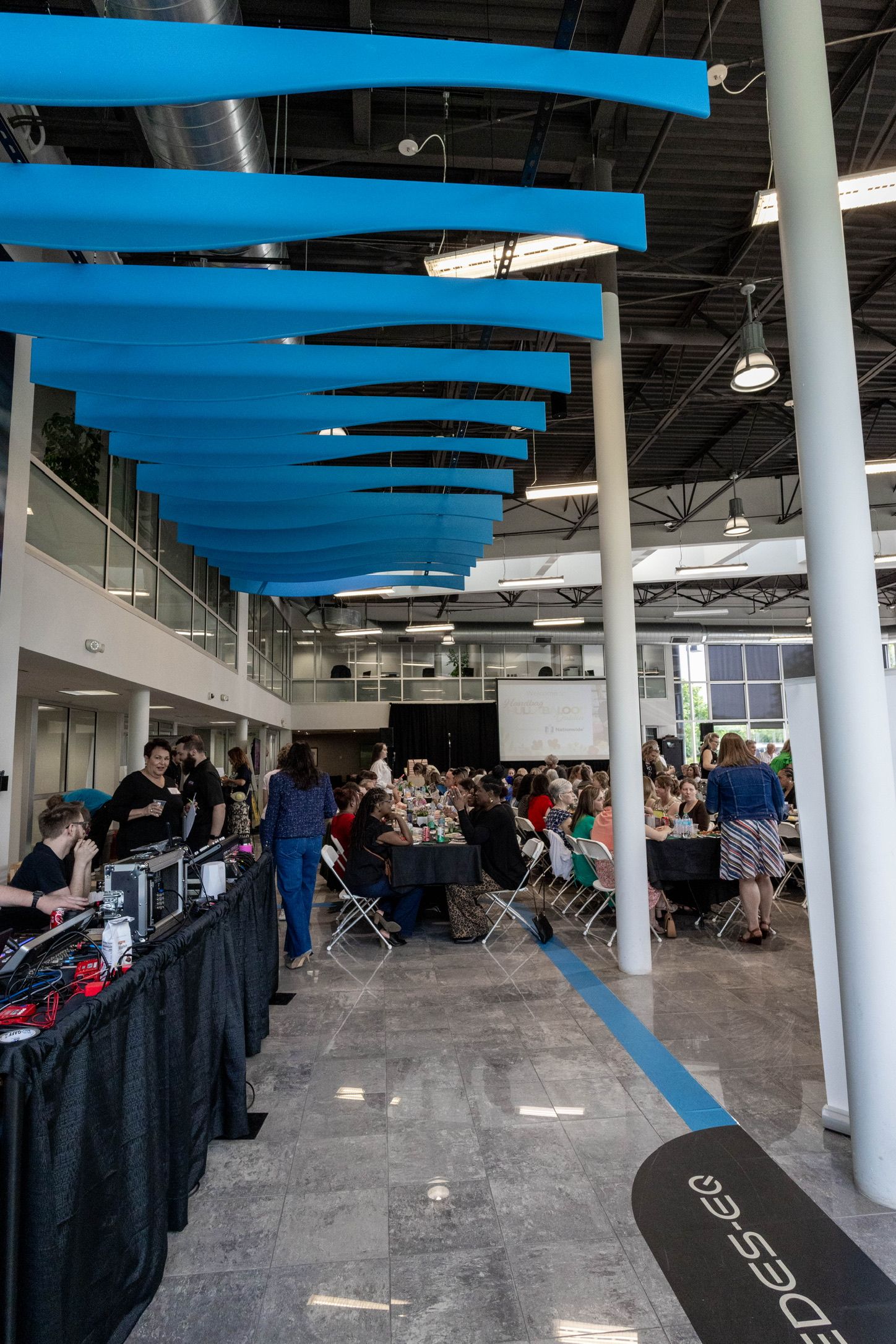 Large modern event space at with distinctive blue ceiling beams hosting a crowded indoor gathering.