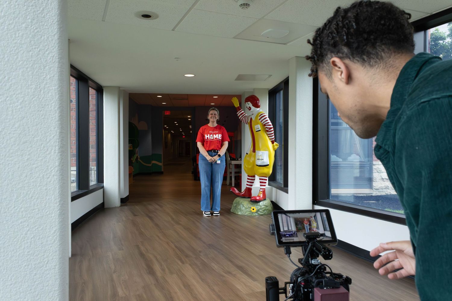Videographer filming a woman in red shirt standing near Ronald McDonald statue in a colorful hallway.