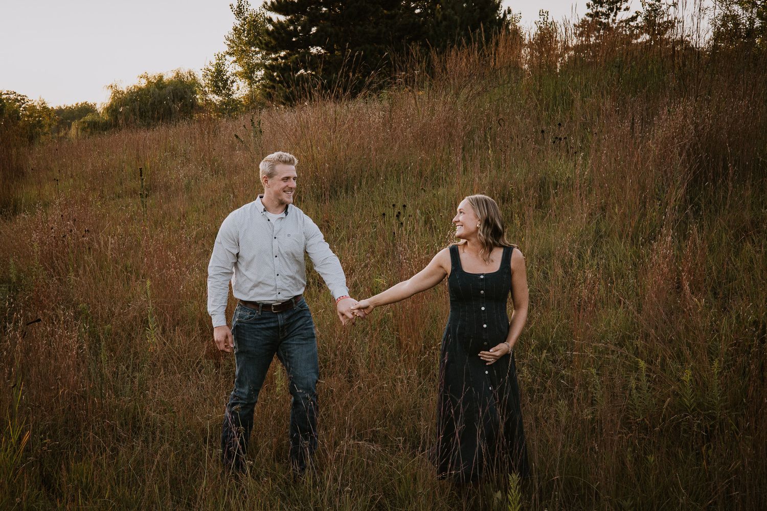 Couple holding hands smiling in a golden field at sunset, wearing casual attire in a romantic outdoor setting.