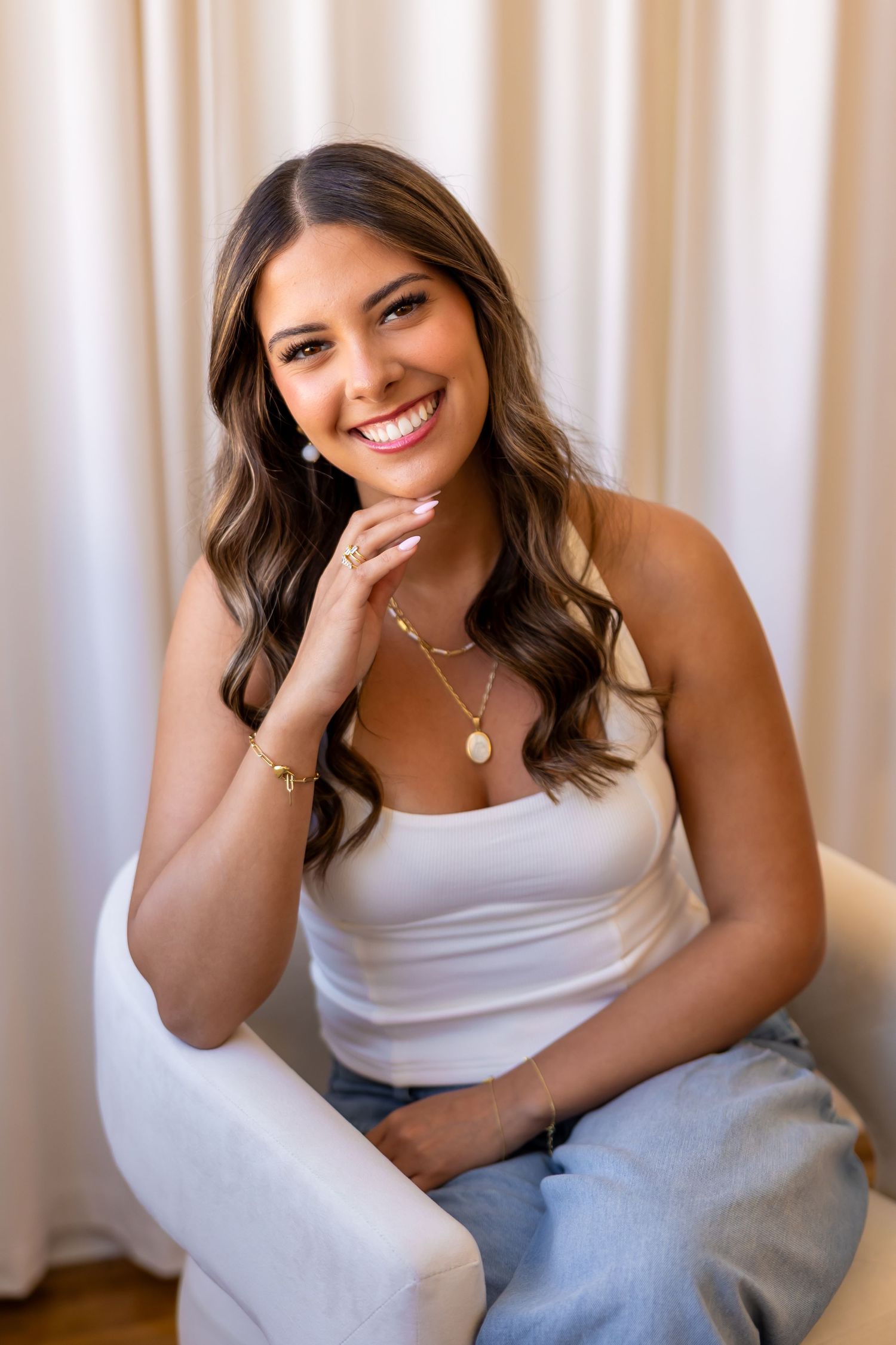 A smiling young woman with long brown hair sits in a chair wearing a white top.